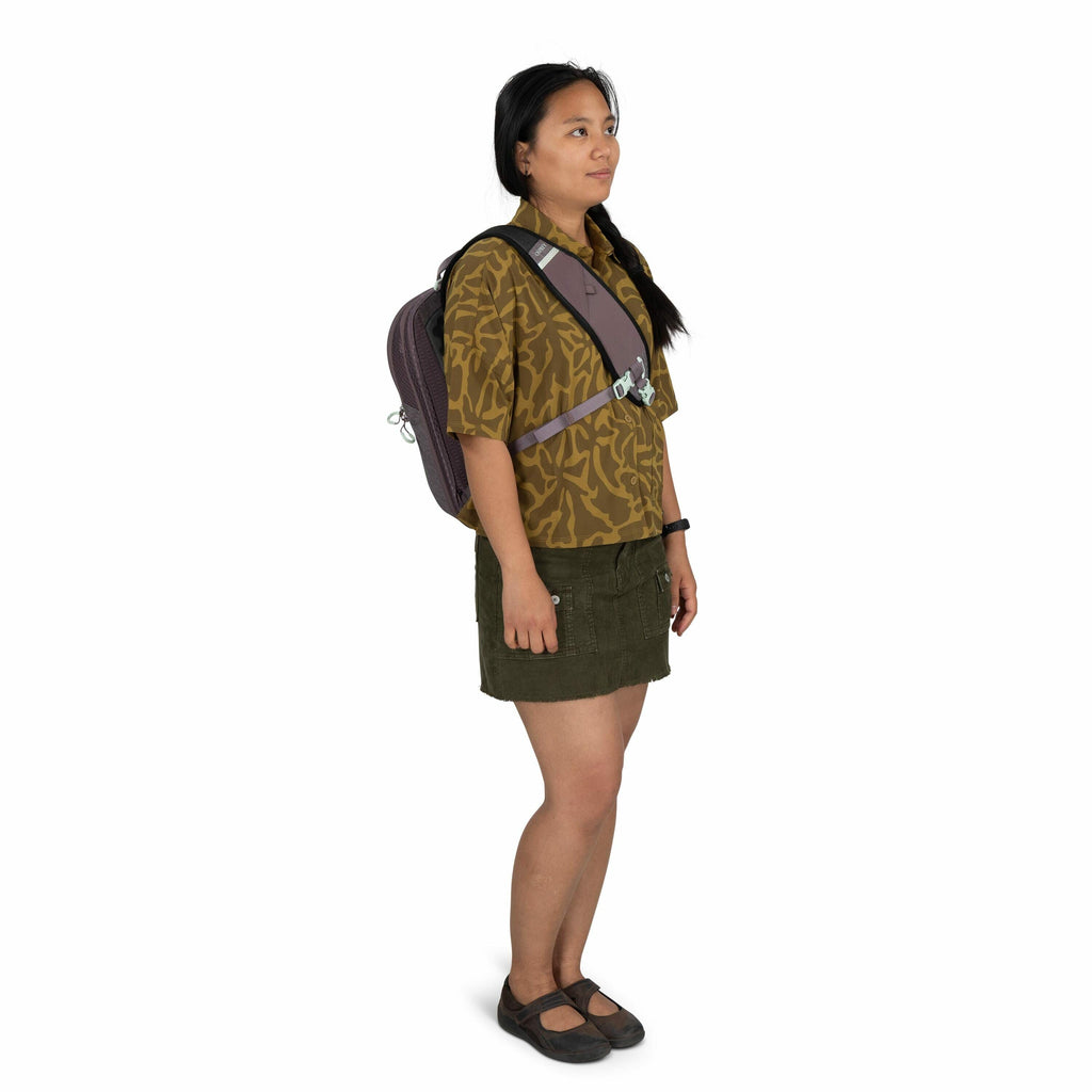 The image depicts a person standing and dressed casually. They are wearing a short-sleeve brown shirt with a yellow abstract pattern, paired with a short green skirt featuring pockets and buttons. On their feet, they have black Mary Jane style shoes. The