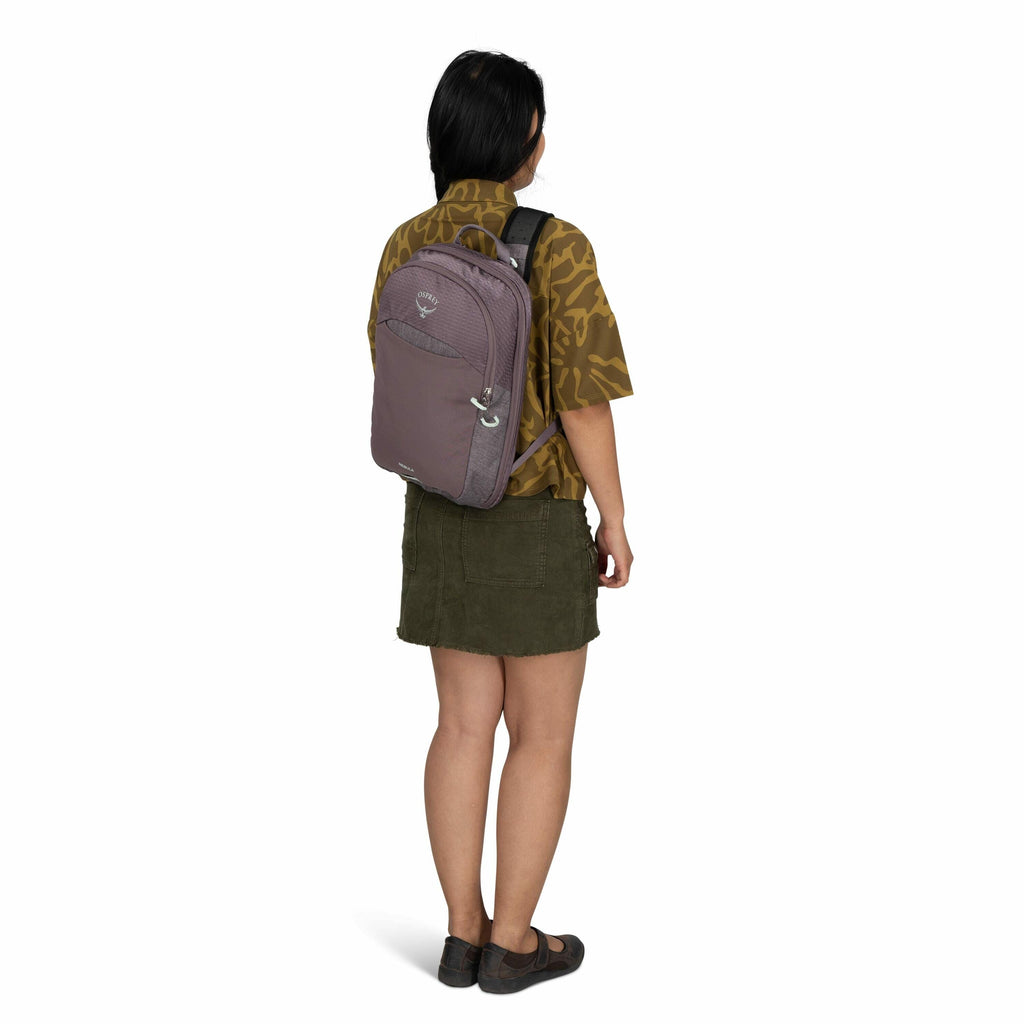 The image features a woman standing with her back facing the camera, wearing a backpack. The backpack is a muted mauve color with the Osprey logo and name visible on the back pocket. She is dressed in an olive green short skirt and a dark green patterned