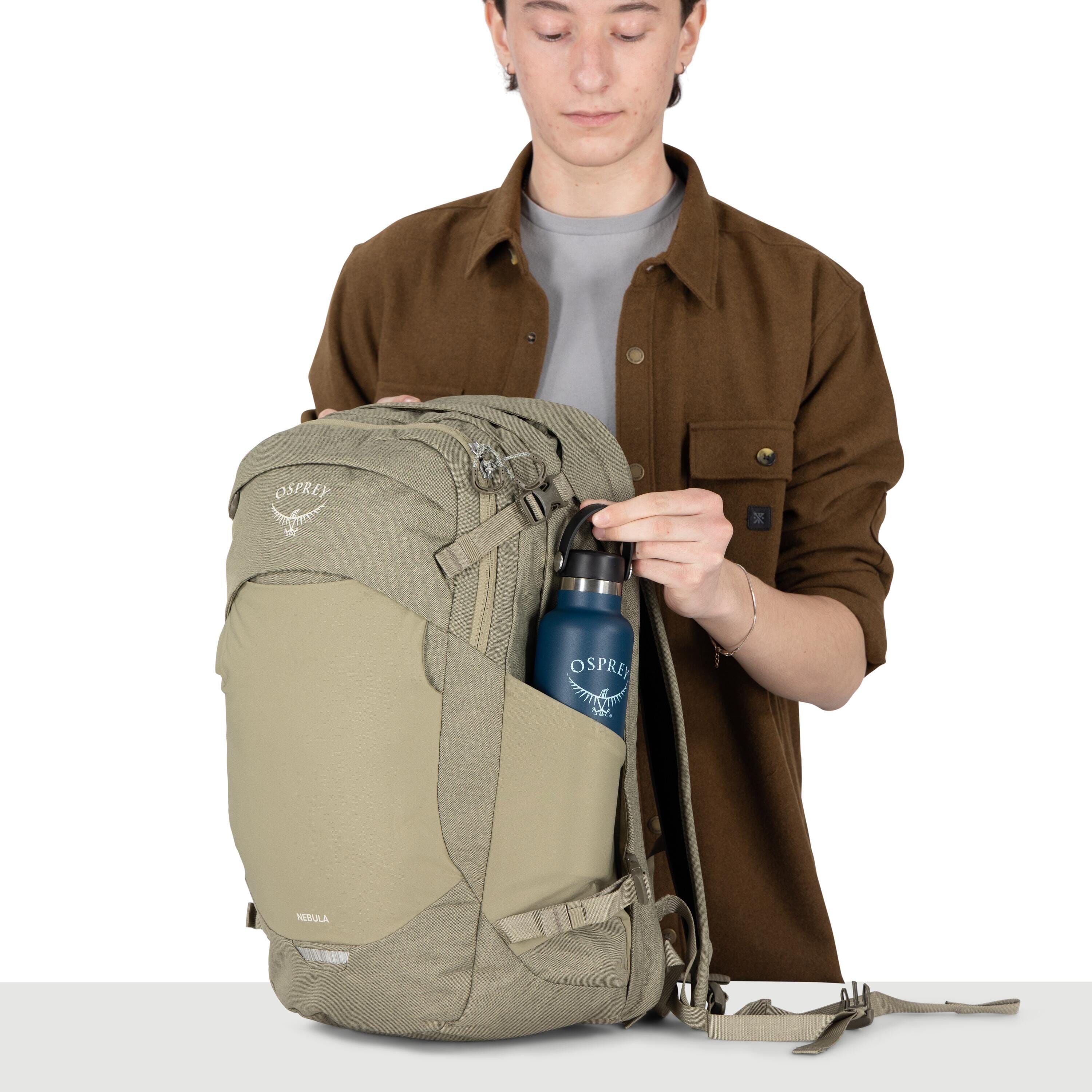 A person is holding a light-olive Osprey backpack with multiple zippered compartments and compression straps. They are placing a blue Osprey-branded water bottle into the stretch side pocket. The person wears a brown overshirt over a gray shirt, and their