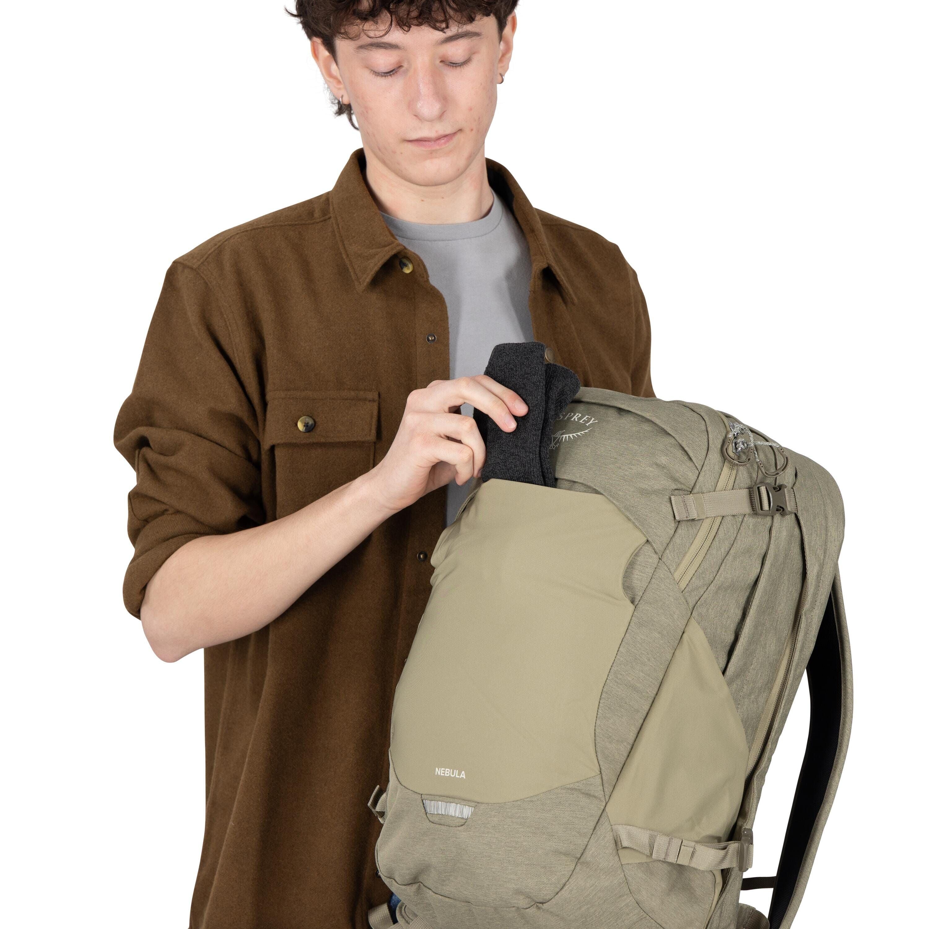 A person is holding a khaki and olive green backpack and placing a dark gray item into the front shove-it pocket. The backpack has compression straps, multiple zippers, and the logo "Osprey" on the upper panel with the model name "Nebula" on the front. Th
