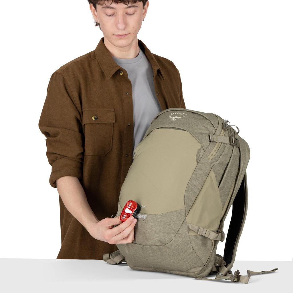 A person stands behind a table holding a gray and khaki Osprey backpack. They are wearing a brown overshirt over a gray tee, with sleeves rolled up. In their hand is a small red keychain or accessory, which they are placing near the front pocket of the ba