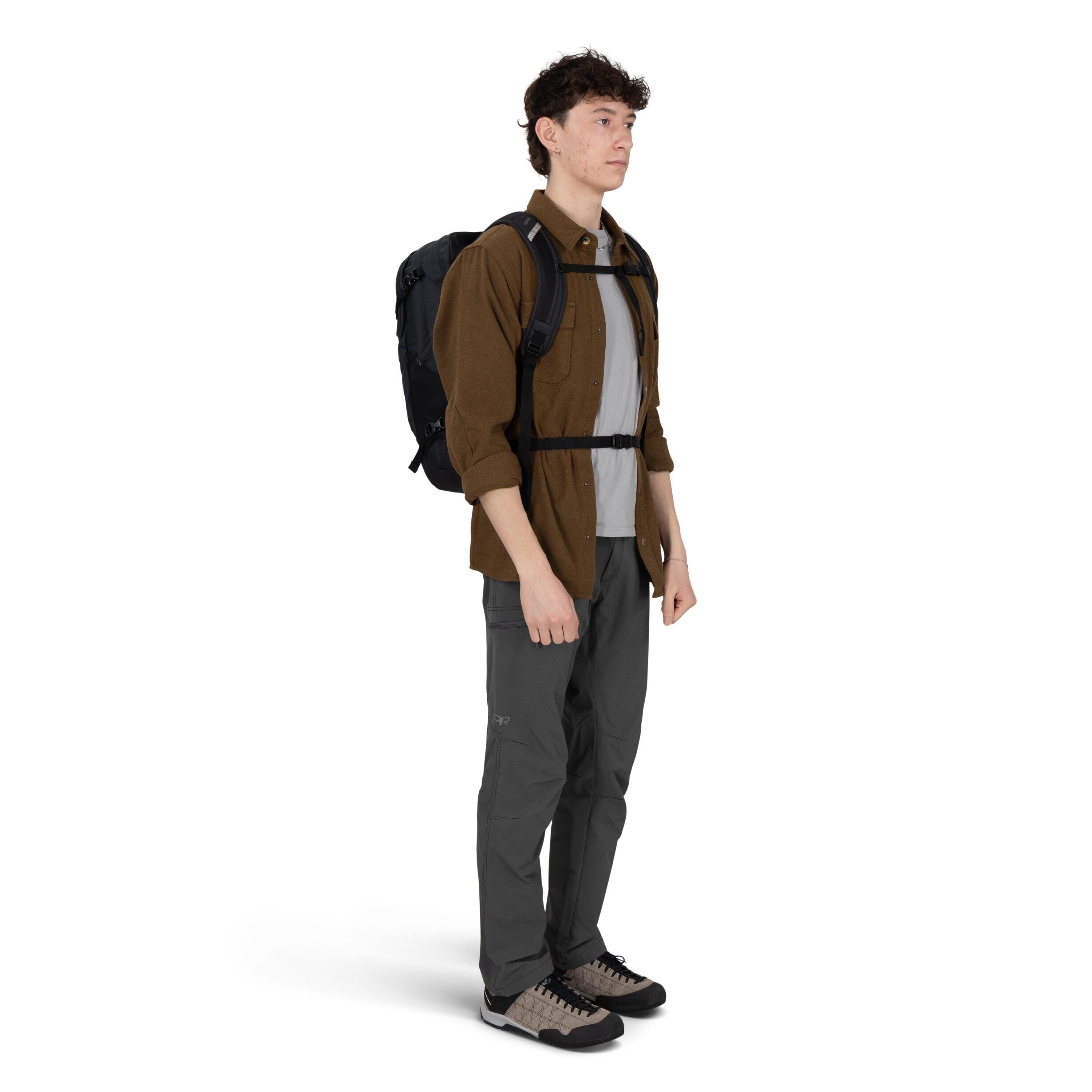 A person stands in a three-quarter view wearing outdoor clothing and a black backpack with chest and waist straps. They have a brown button-up shirt over a light gray T-shirt, gray technical pants with articulated knees, and beige-and-black hiking shoes.