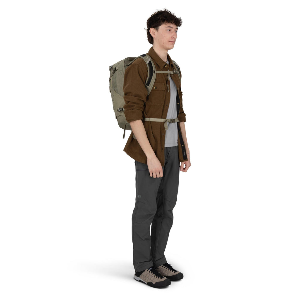 A person is standing in a three-quarter pose wearing outdoor clothing and a backpack. They have on a brown button-up overshirt over a light gray T-shirt, charcoal-gray hiking pants, and beige-and-black approach-style shoes. The backpack is olive green wit