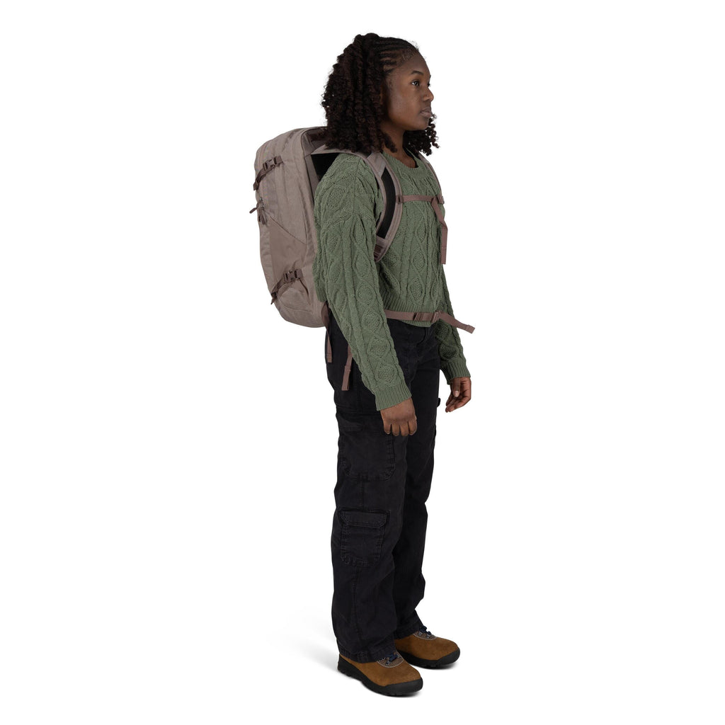 A person stands in profile wearing outdoor clothing and a large beige backpack with chest and waist straps. They have on a green cable_knit sweater, black cargo pants, and brown hiking shoes with black soles. The backpack features side compression straps