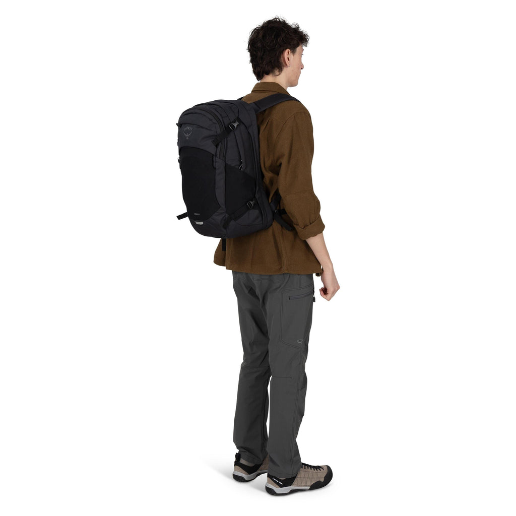 A person wearing outdoor clothing stands in a three-quarter rear view with a dark gray and black Osprey backpack on their back. They have on a brown long-sleeve shirt, gray technical pants with zip pockets and knee panels, and beige-and-black hiking shoes