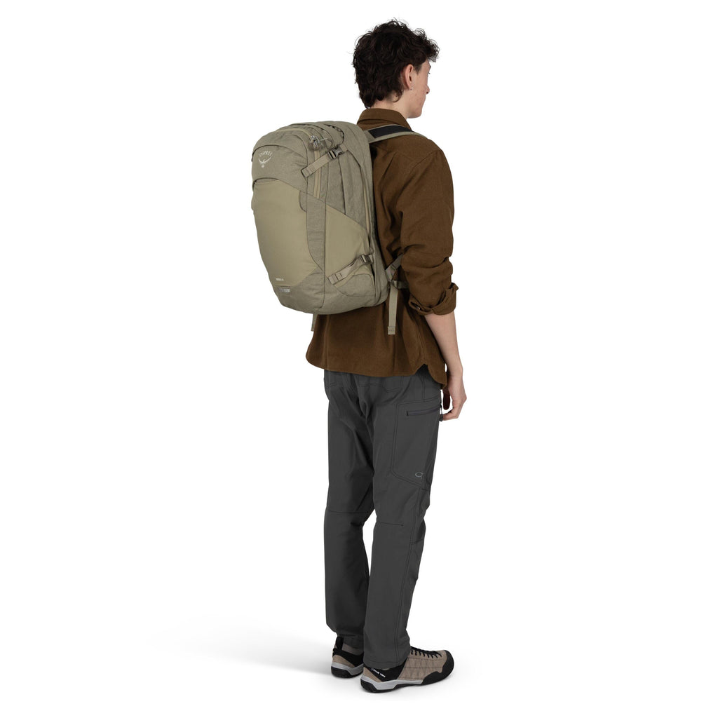 A person is standing in a side-back view wearing an olive green Osprey backpack. They're dressed in a brown long-sleeve shirt, dark gray hiking pants with articulated knees, and tan-and-black approach-style shoes. The backpack has multiple compression str