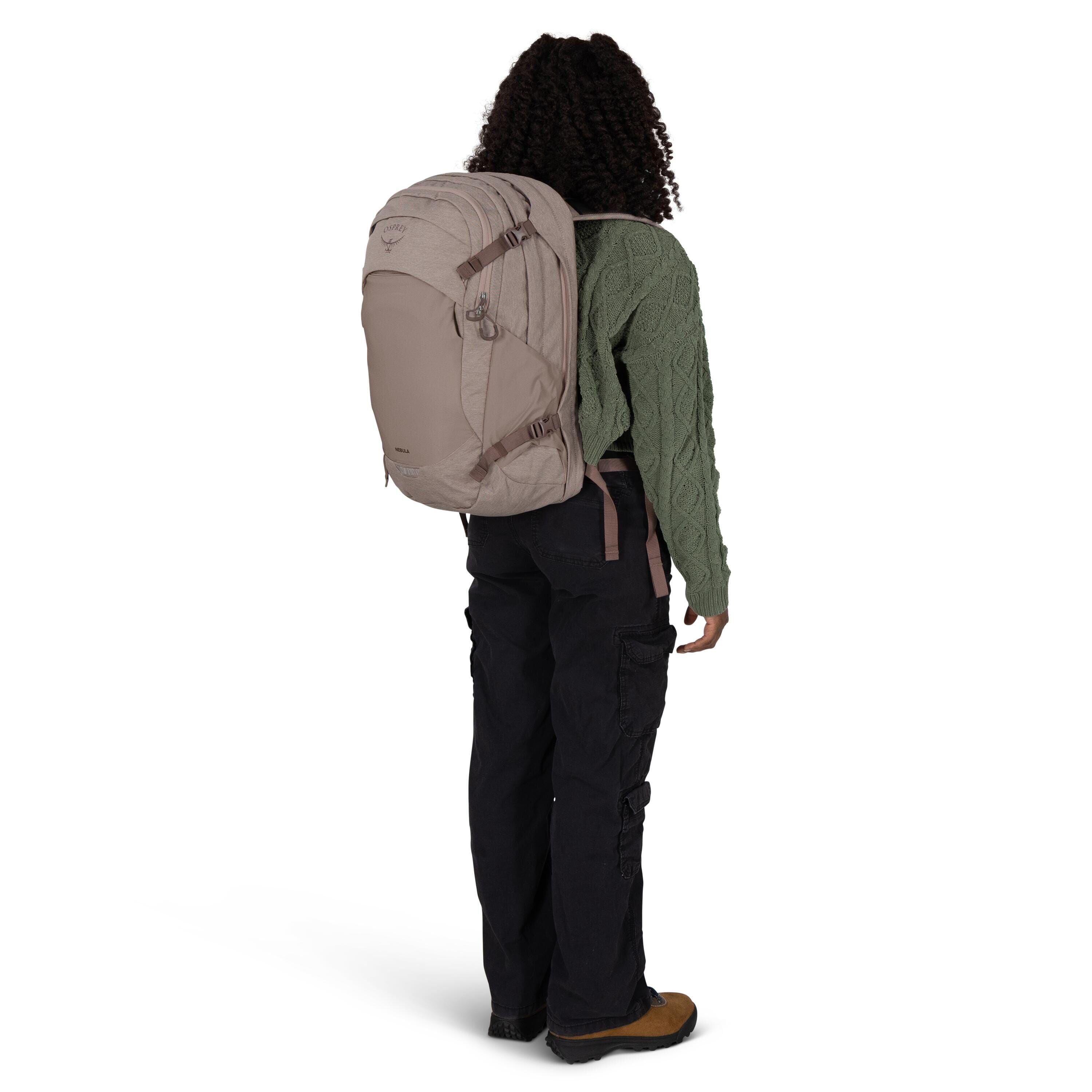A person is shown from the back wearing a large, taupe-colored Osprey backpack. They are dressed in a green cable-knit sweater, black cargo pants, and brown hiking shoes with rugged soles. The backpack has multiple compartments, compression straps, and a