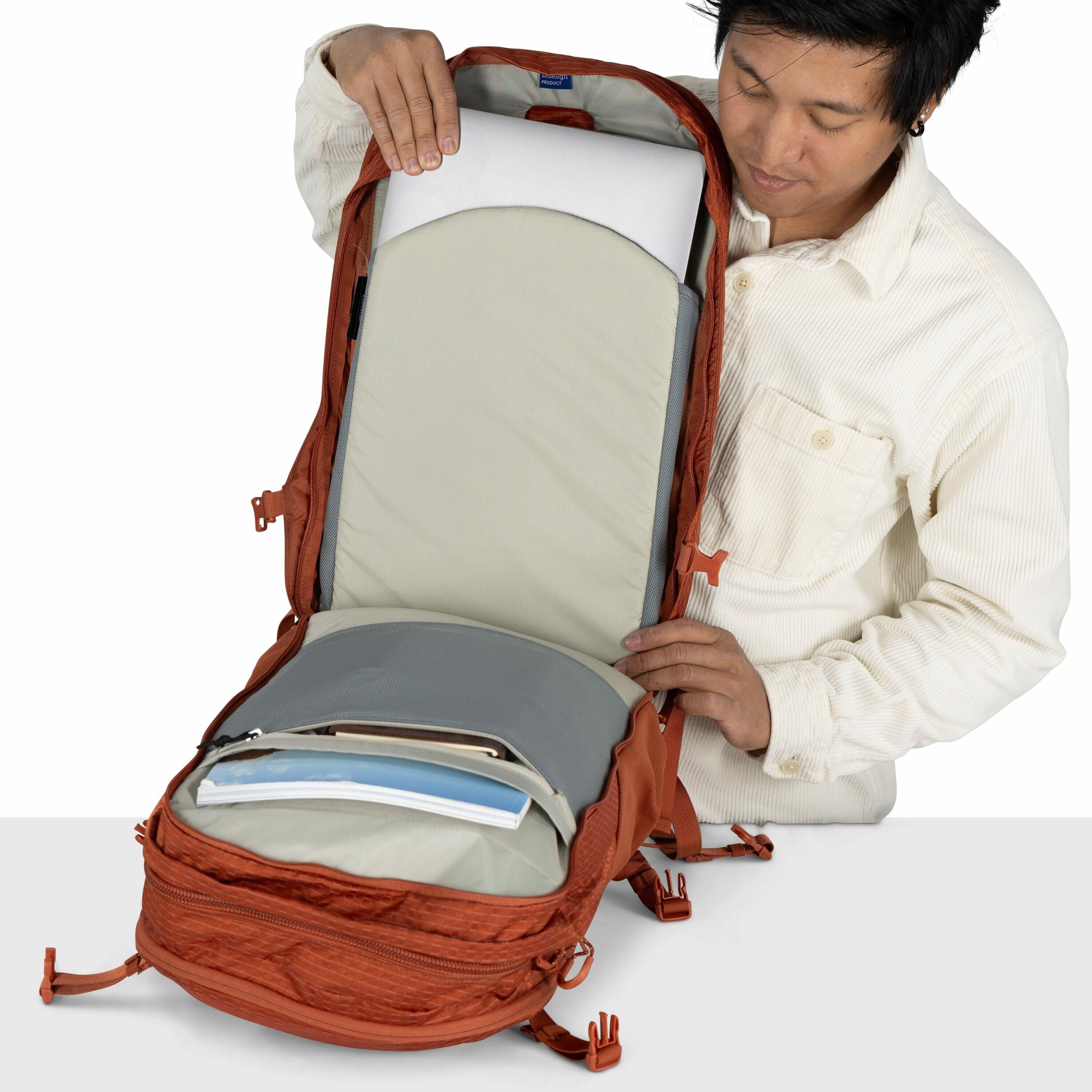 The image shows a person opening an orange backpack, revealing its spacious interior. Inside the backpack, a padded compartment holds a laptop, and in front of this, there is a large, shallow compartment with documents or folders. The interior of the back