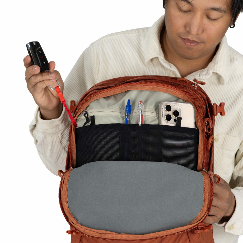 In the image, a person is holding open an orange backpack to reveal its organized interior. The backpack features several pockets that hold various items, including a blue pen, a red pen, and a smartphone with a triple-camera setup. The individual is hold