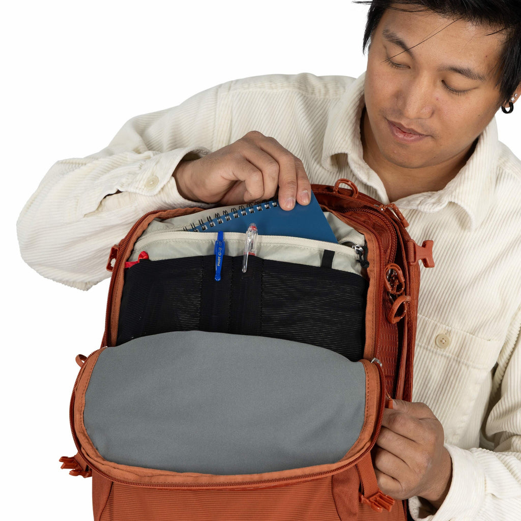 The image shows a person opening a burnt-orange colored backpack to reveal its contents. Inside the backpack, there is a blue spiral notebook being inserted or taken out by the person's right hand. Additionally, the backpack contains a few pens, including