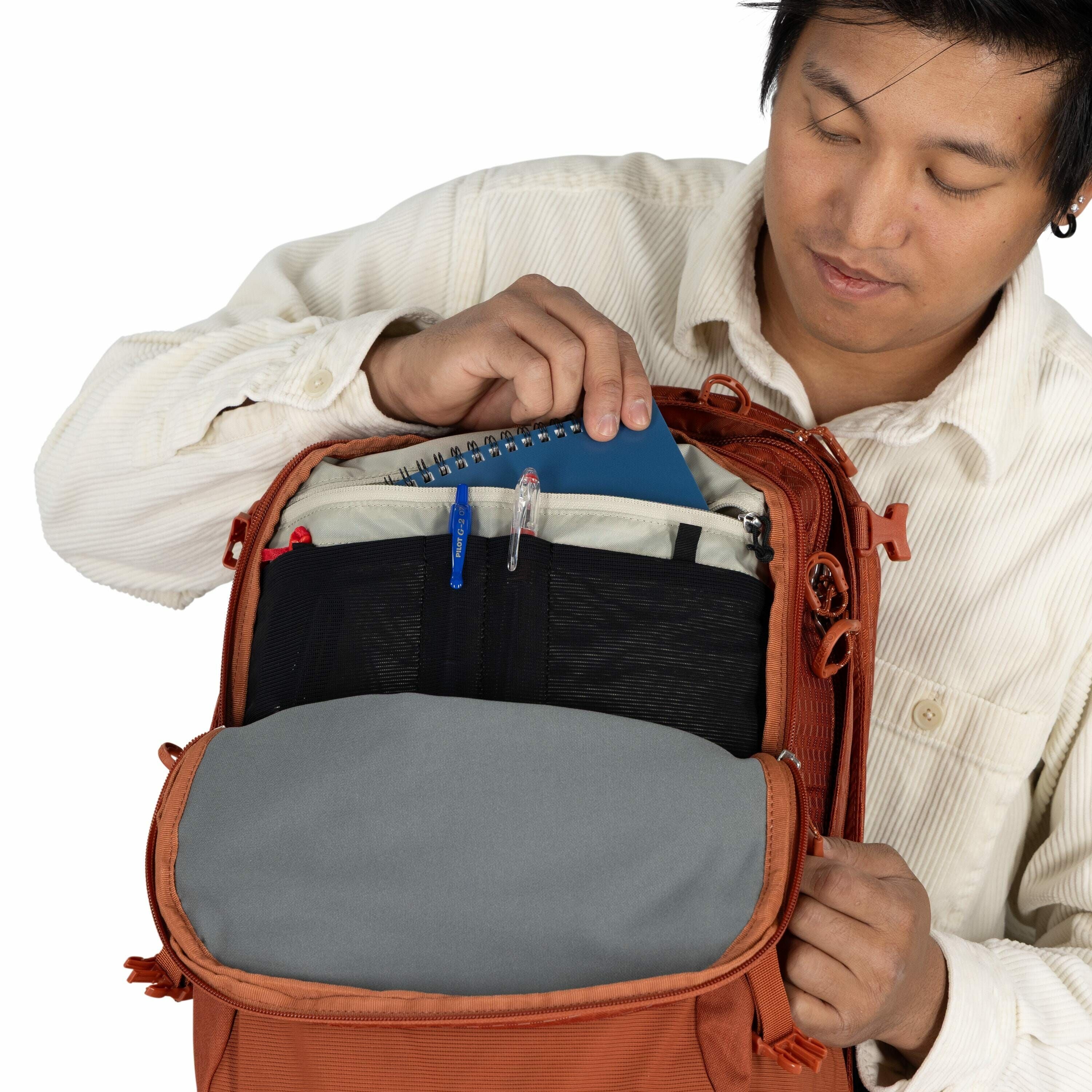 The image shows a person opening a burnt-orange colored backpack to reveal its contents. Inside the backpack, there is a blue spiral notebook being inserted or taken out by the person's right hand. Additionally, the backpack contains a few pens, including