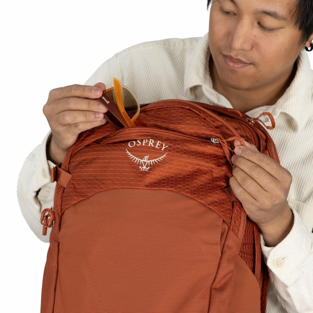 The image shows a person holding an orange backpack made by Osprey. The backpack features a textured fabric design with the Osprey logo, which includes the brand name and a stylized bird graphic, prominently displayed on the front pocket. The person is un