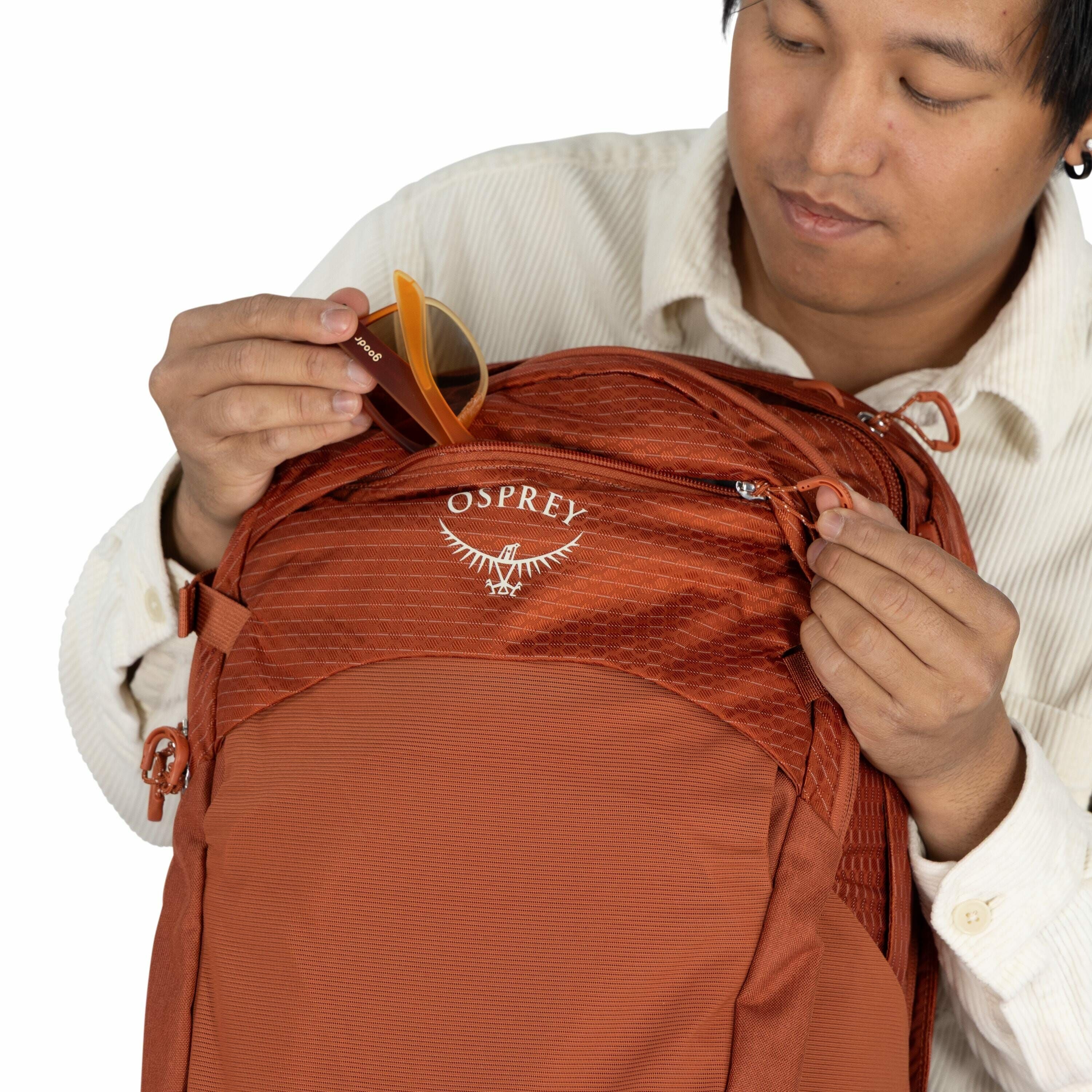 The image shows a person holding an orange backpack made by Osprey. The backpack features a textured fabric design with the Osprey logo, which includes the brand name and a stylized bird graphic, prominently displayed on the front pocket. The person is un
