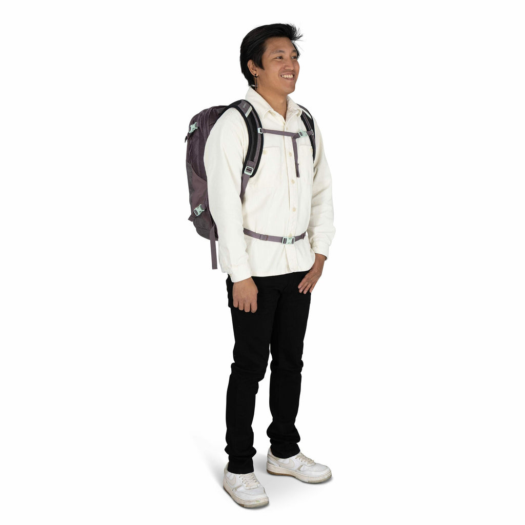 The image shows a person standing and dressed casually, with a purple backpack strapped securely. They are wearing a cream-colored button-up shirt and black pants, paired with white sneakers that have noticeable wear marks. The backpack features black str