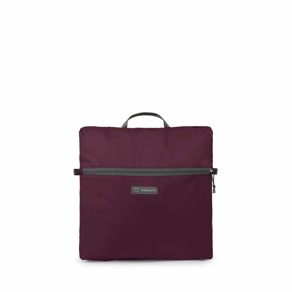 The images display a foldable maroon backpack with a simple and compact design. The backpack is shown in a folded state, with the main rectangular body and a front zipper pocket visible. It features a carrying handle at the top, and the texture of the mat