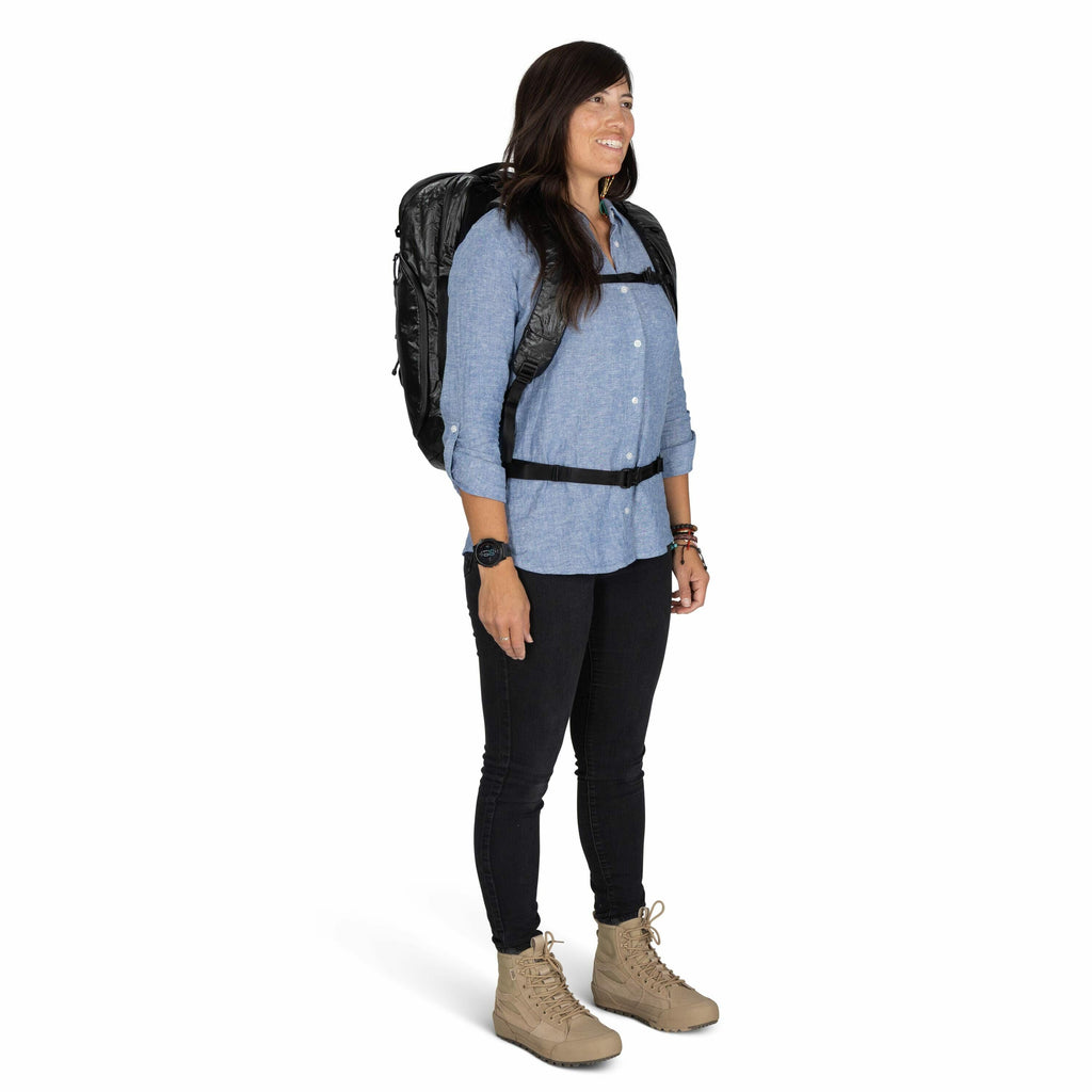 The image shows a person standing, dressed in casual outdoor attire. They are wearing a blue button-up shirt paired with black pants and tan lace-up boots. The person is carrying a black backpack secured with chest and waist straps, indicating preparednes