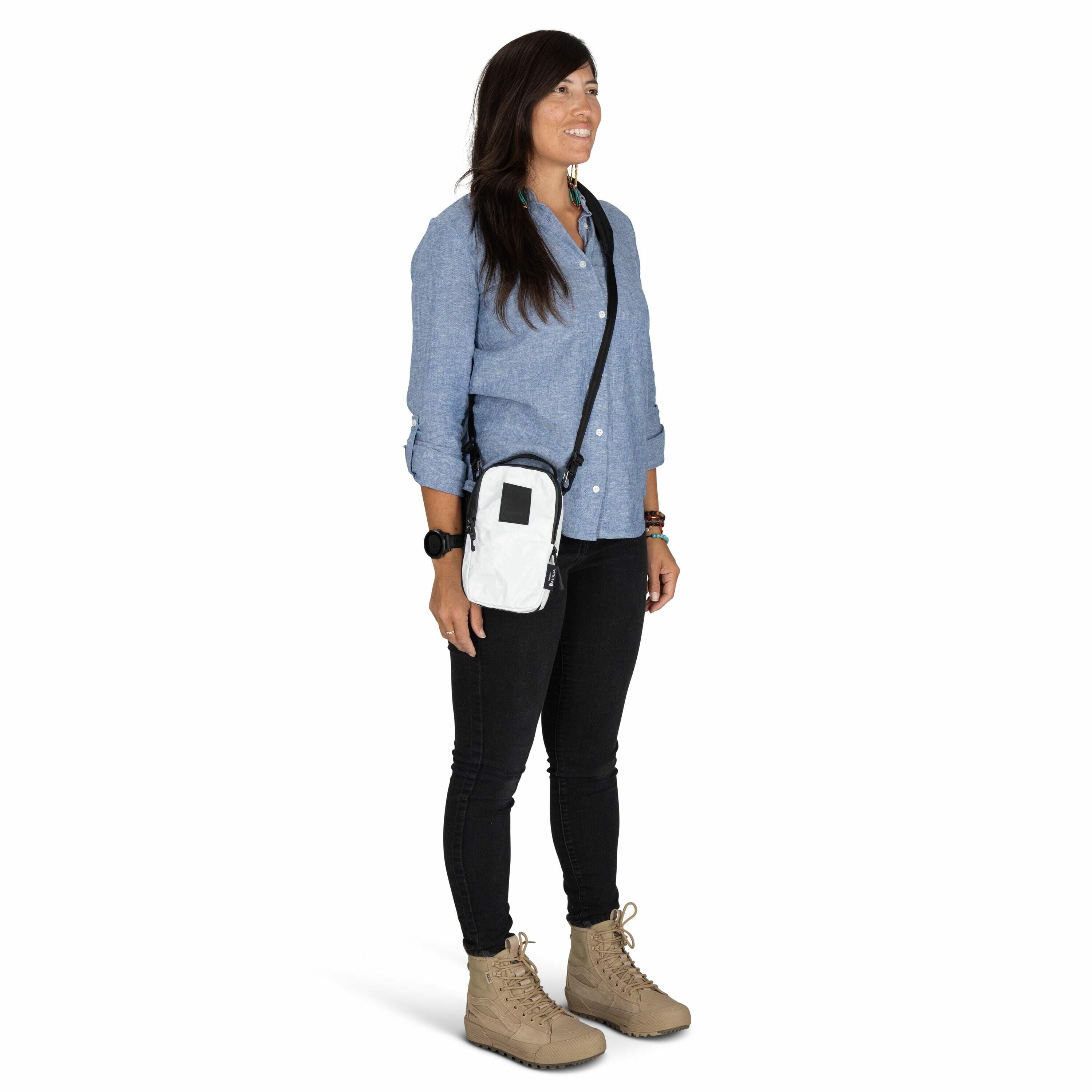 The image shows a person standing against a plain white background. They are wearing a light blue button-up shirt with the sleeves rolled up, black pants, and beige lace-up ankle boots. A white crossbody bag with a black strap is slung across their should