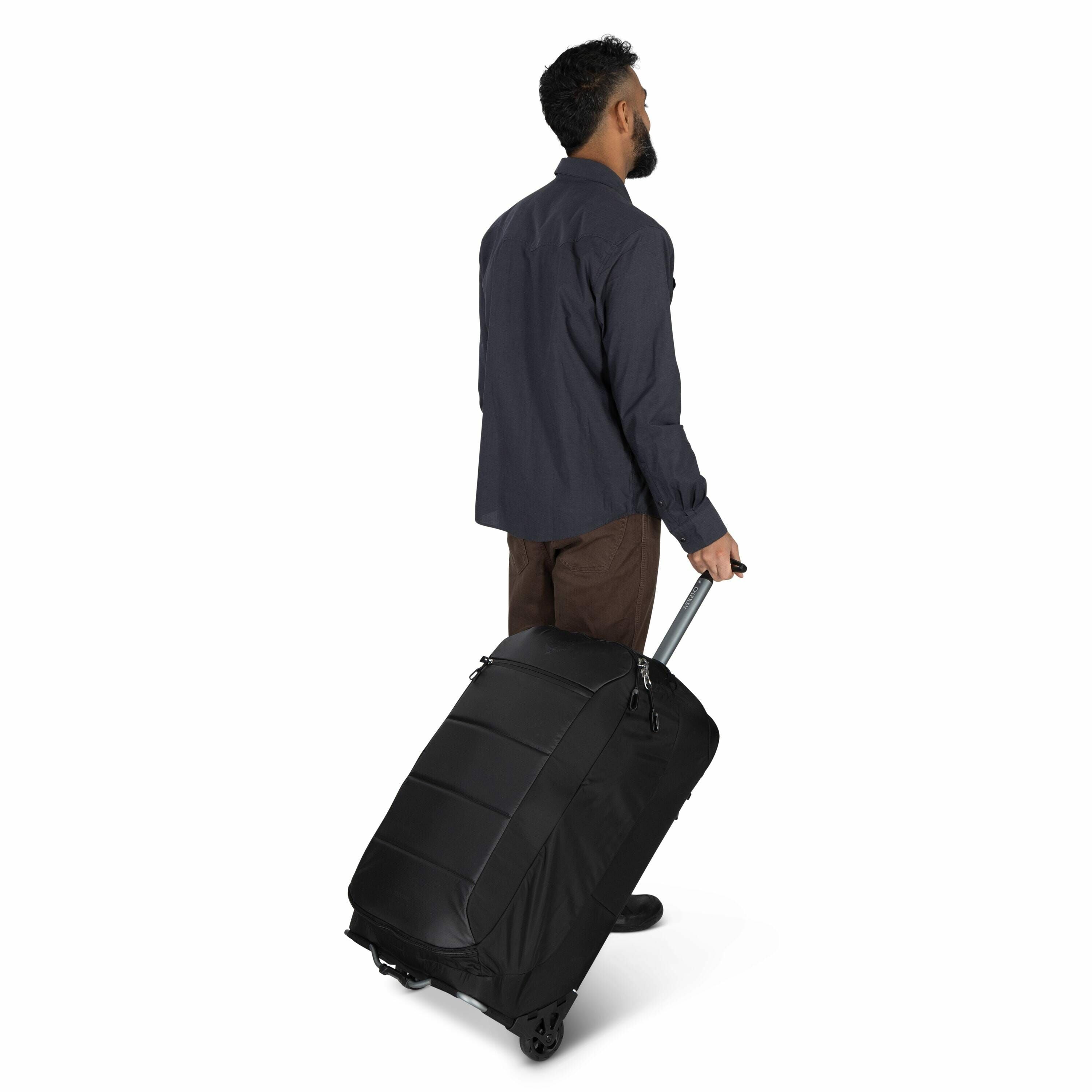 The image shows a man with a short beard, viewed from behind, walking away while pulling a large black wheeled suitcase with one hand. He is wearing a dark blue button-up shirt, brown trousers, and black shoes. The suitcase is equipped with sturdy wheels
