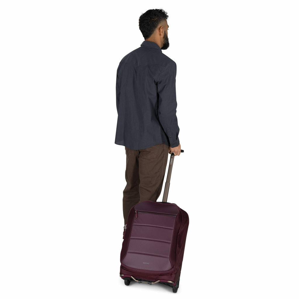 The image shows a man with dark hair and a beard standing and facing away from the camera. He is wearing a dark blue button-up shirt and brown pants. The man is pulling a burgundy wheeled suitcase, which features horizontal ridges on its surface, using th
