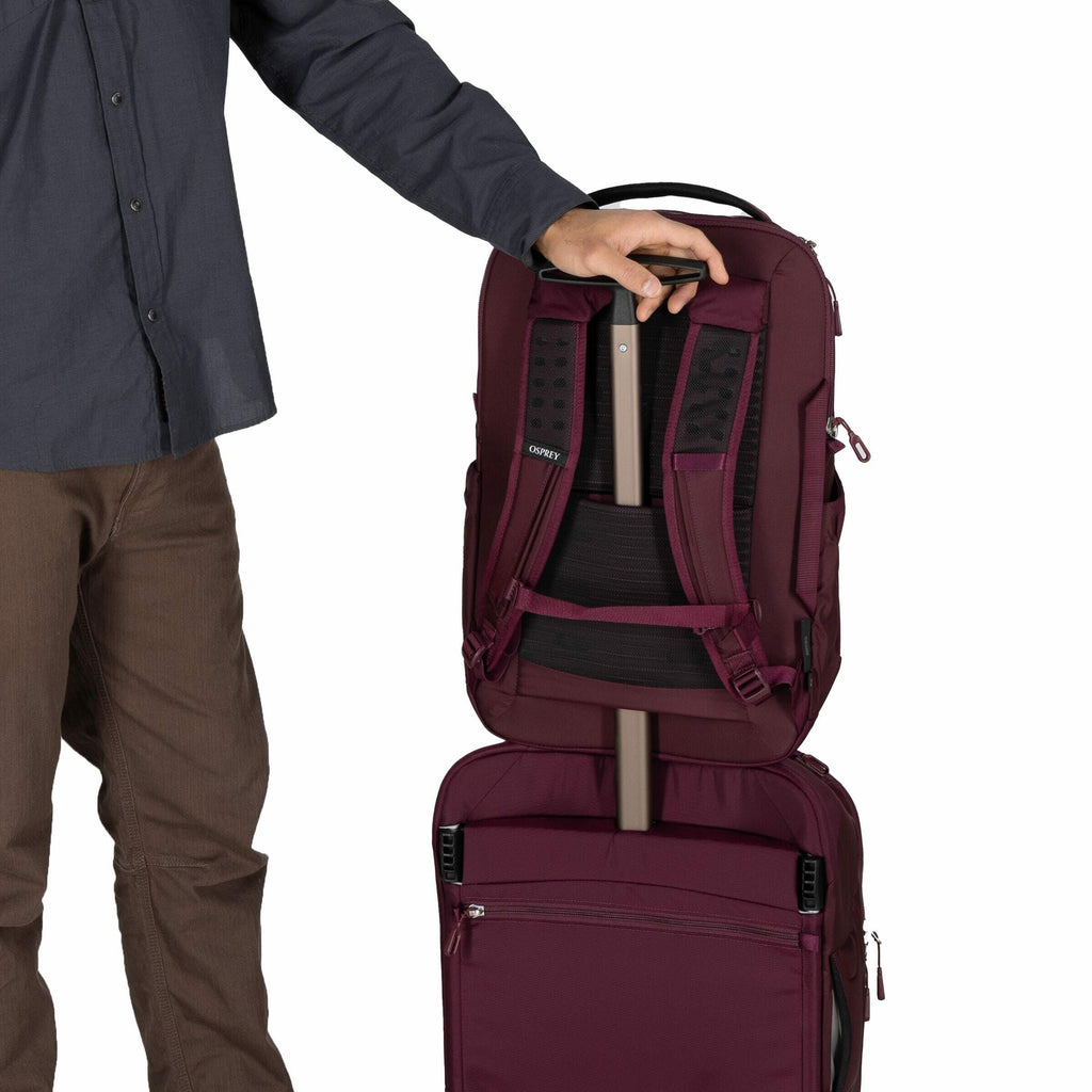 The image shows a person standing beside a luggage setup, with only their torso and legs visible, wearing brown pants and a dark gray button-up shirt. They are holding a maroon backpack, which is being slipped over the extended handle of a rolling suitcas
