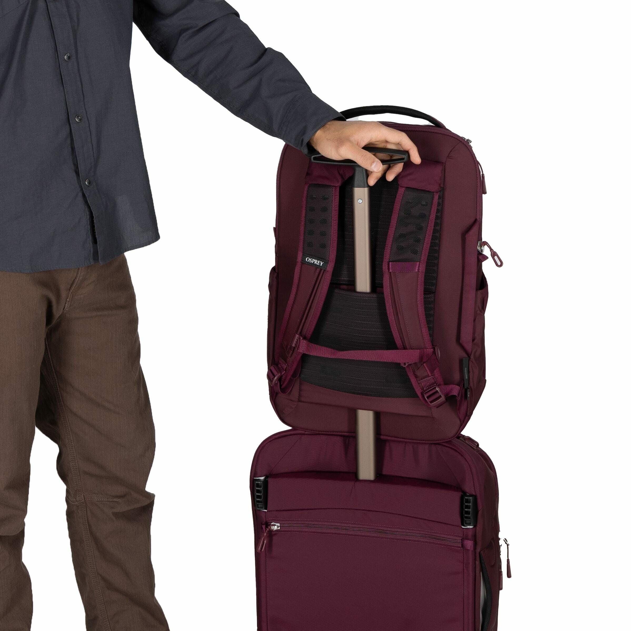 The image shows a person standing beside a luggage setup, with only their torso and legs visible, wearing brown pants and a dark gray button-up shirt. They are holding a maroon backpack, which is being slipped over the extended handle of a rolling suitcas