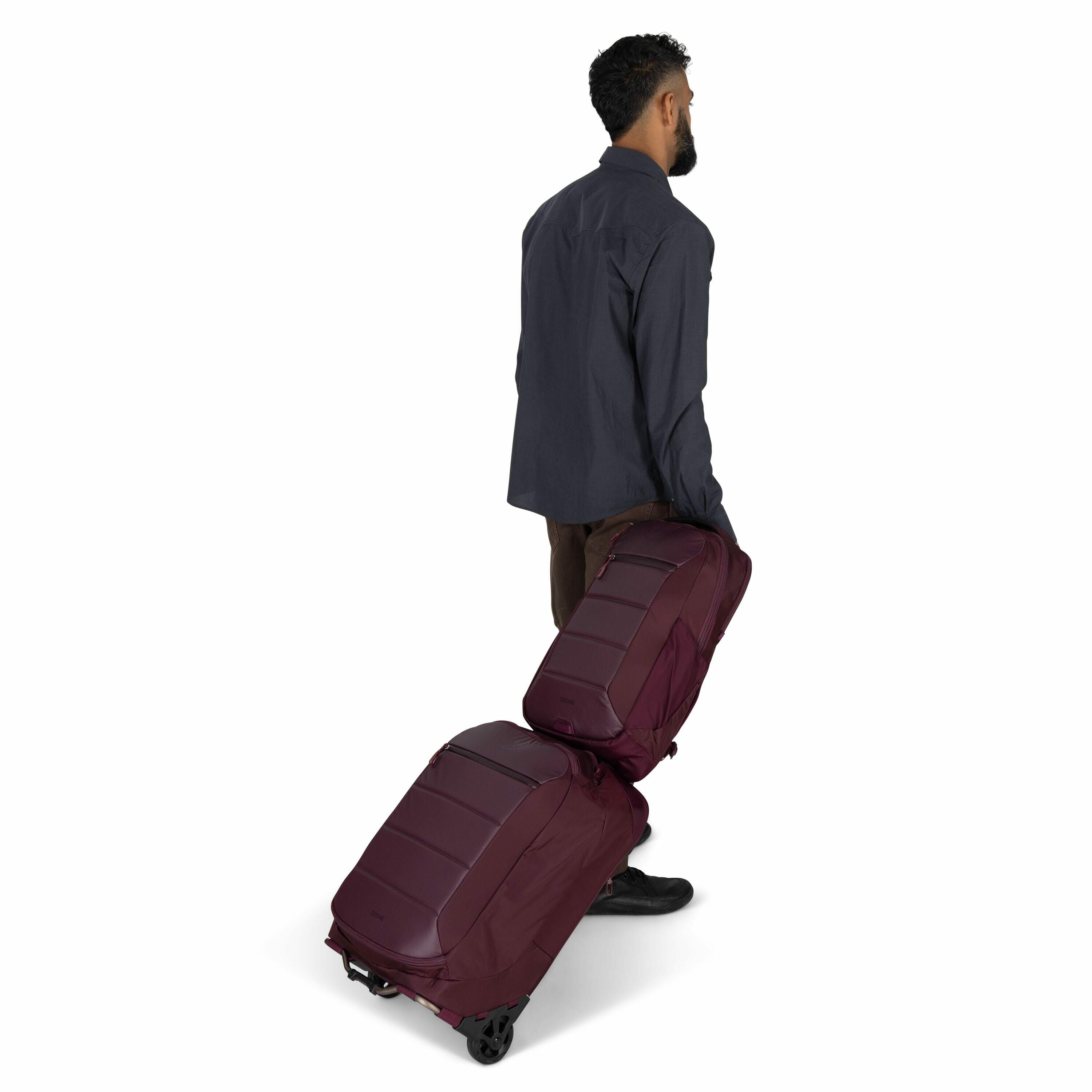 The image shows a man with dark hair and a beard, wearing a dark gray shirt and brown pants, walking while pulling two maroon-colored wheeled luggage bags. The luggage appears to be linked together, with one suitcase trailing behind the other. The man is