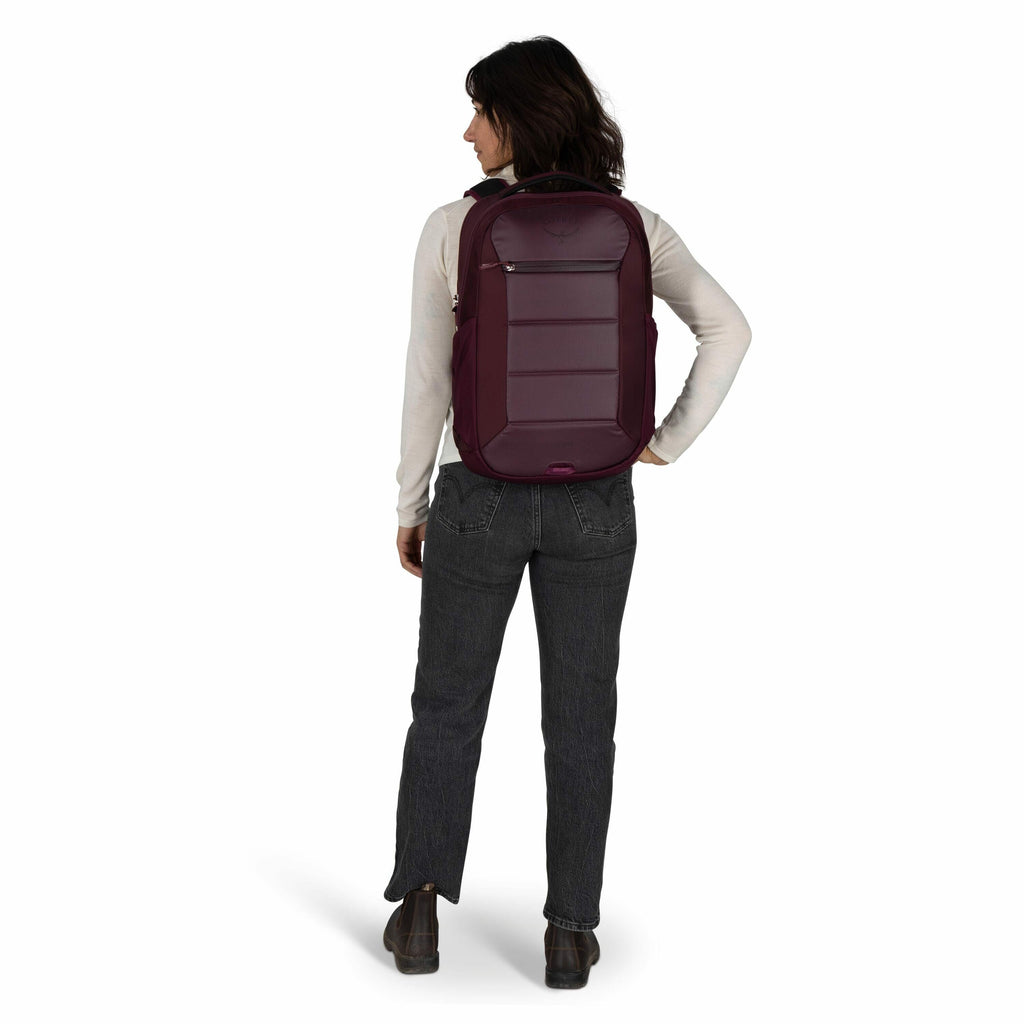 The image shows a person standing with their back facing the camera, wearing a dark burgundy backpack. The individual is dressed in a light-colored long-sleeve top and dark gray jeans, paired with dark-colored shoes. The backpack appears to have a sleek d