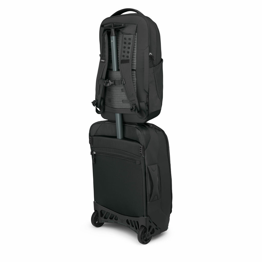 The image shows a black backpack securely attached to the handle of a rolling suitcase. The backpack has a sleek design with multiple compartments and is oriented vertically with the straps and back padding visible. The rolling suitcase below it is also b