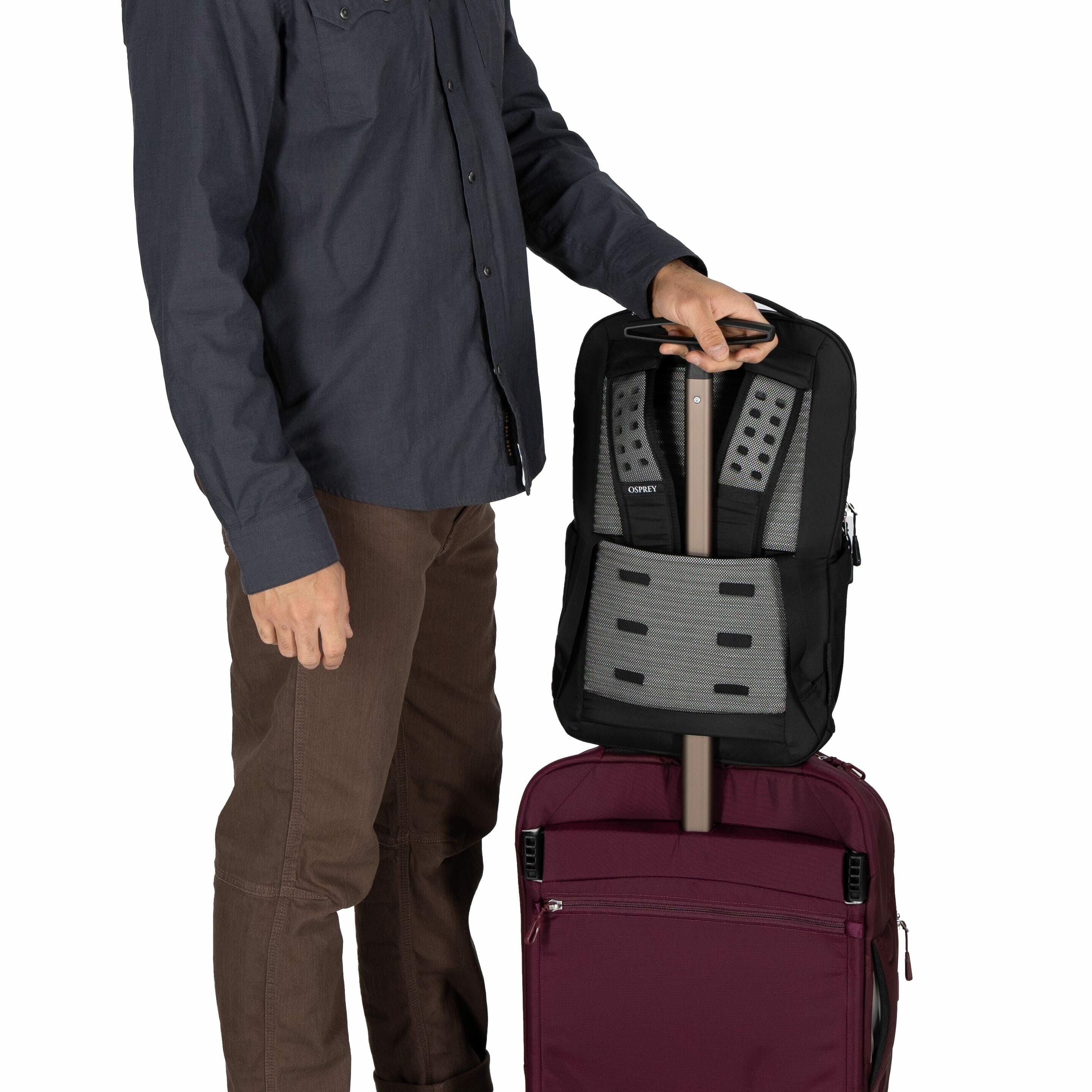 The image shows a person standing next to a maroon suitcase with a telescopic handle extended upwards. The person, dressed in a dark blue shirt and brown pants, is holding a black backpack with a grey padded back panel. The backpack is slipped over the ha