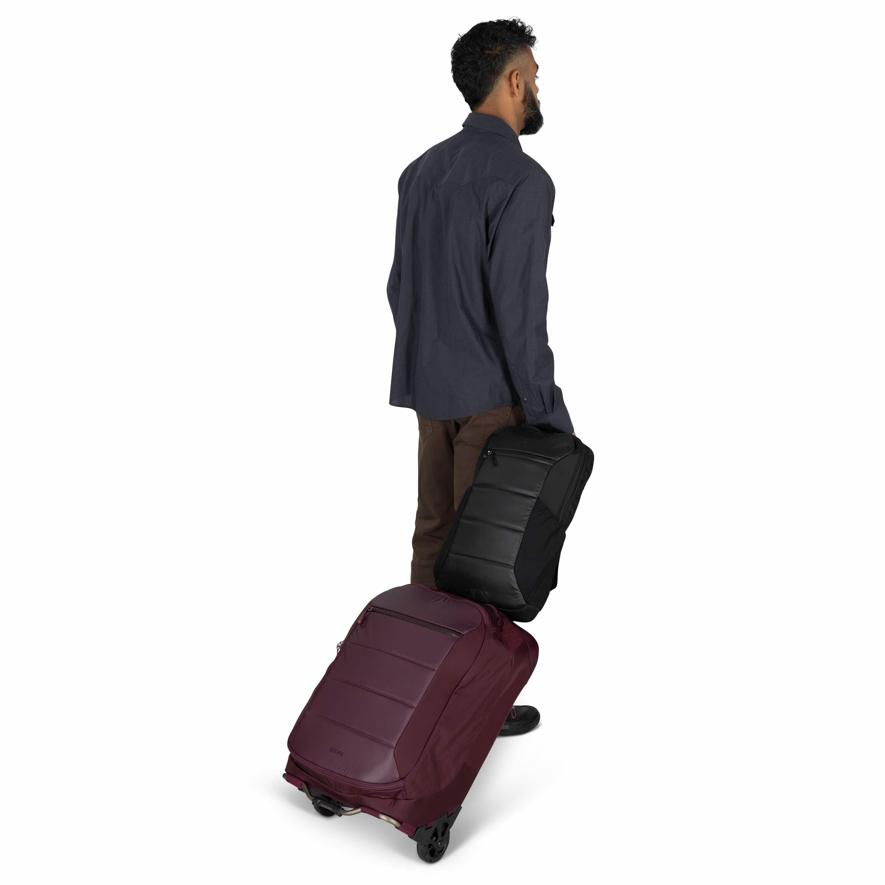 The image shows a person walking away with two pieces of luggage on wheels. The individual is wearing a dark blue button-up shirt and brown pants. One suitcase is a deep burgundy color, and the other is black, with both appearing sleek and modern. The per