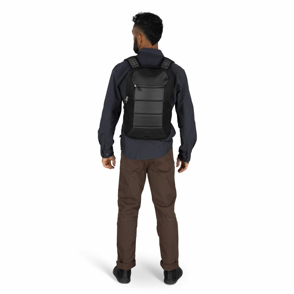 The image shows a man standing with his back facing the camera, wearing a black backpack. The backpack has a sleek design with a hard shell and visible seams, giving it a modern, structured appearance. He is dressed in a dark blue long-sleeve shirt and br