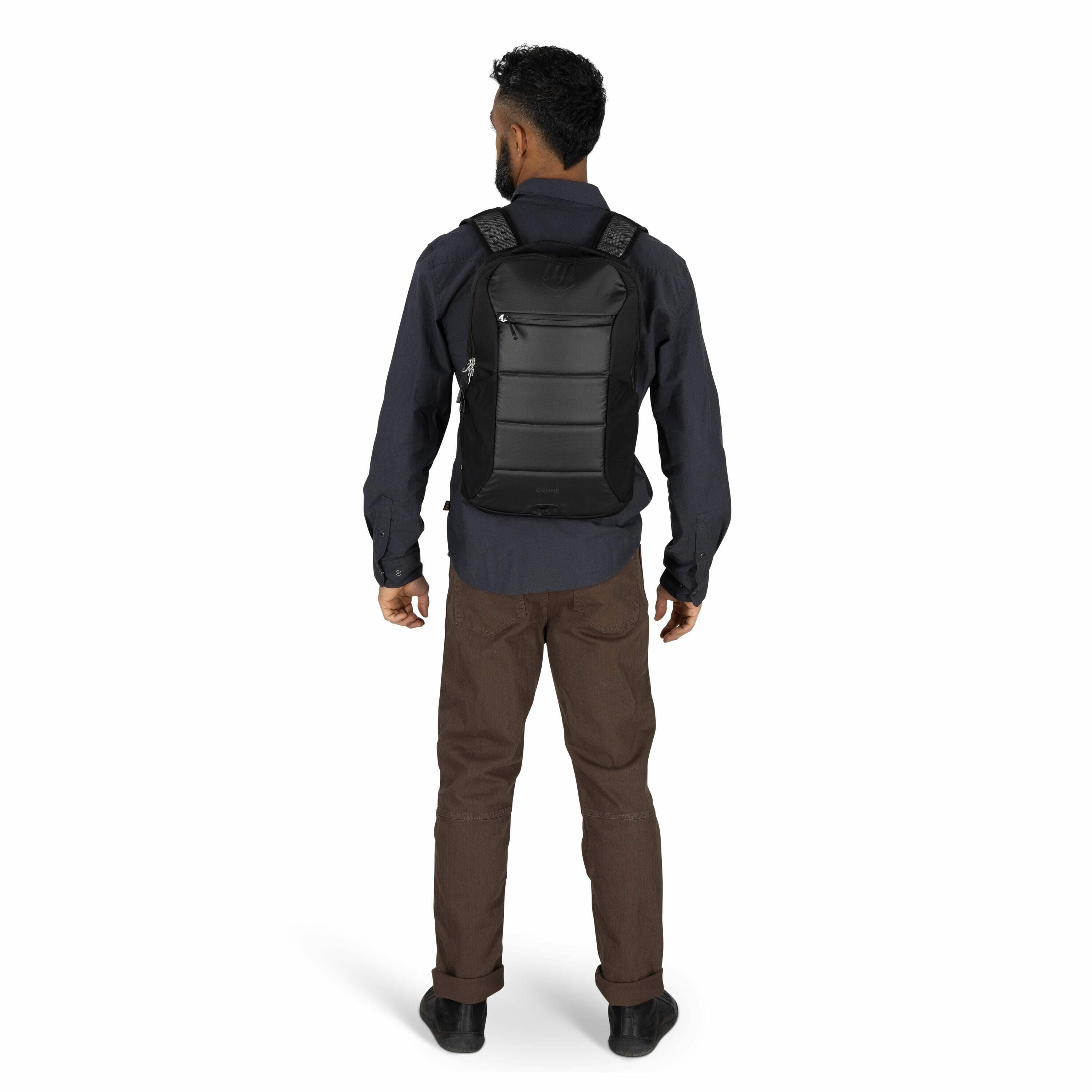 The image shows a man standing with his back facing the camera, wearing a black backpack. The backpack has a sleek design with a hard shell and visible seams, giving it a modern, structured appearance. He is dressed in a dark blue long-sleeve shirt and br