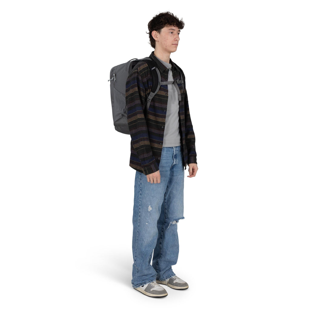 The image shows a person standing and wearing a casual outfit, including a striped button-up shirt layered over a gray t-shirt, loose-fitting distressed blue jeans, and worn sneakers. They have a large gray backpack with chest straps on their back, sugges