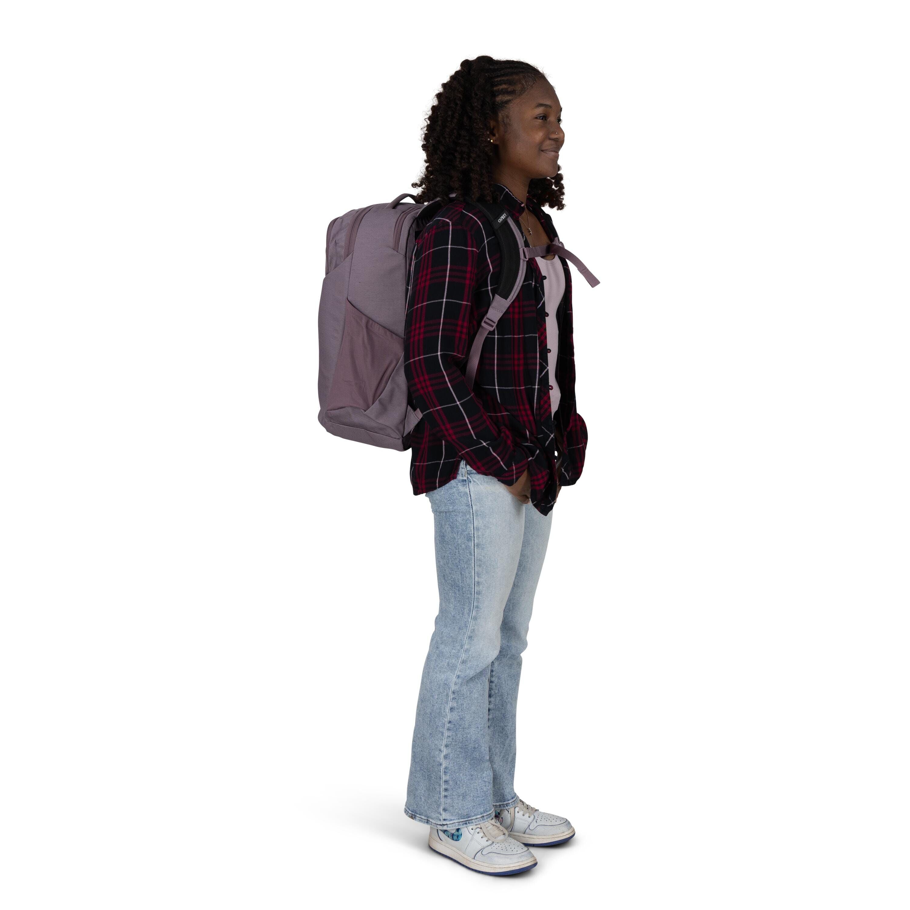 The person in the image is standing and appears to be ready for travel or school, wearing a large purple backpack. They are dressed in a red and black plaid shirt layered over a light shirt, paired with light blue jeans and white sneakers featuring blue d