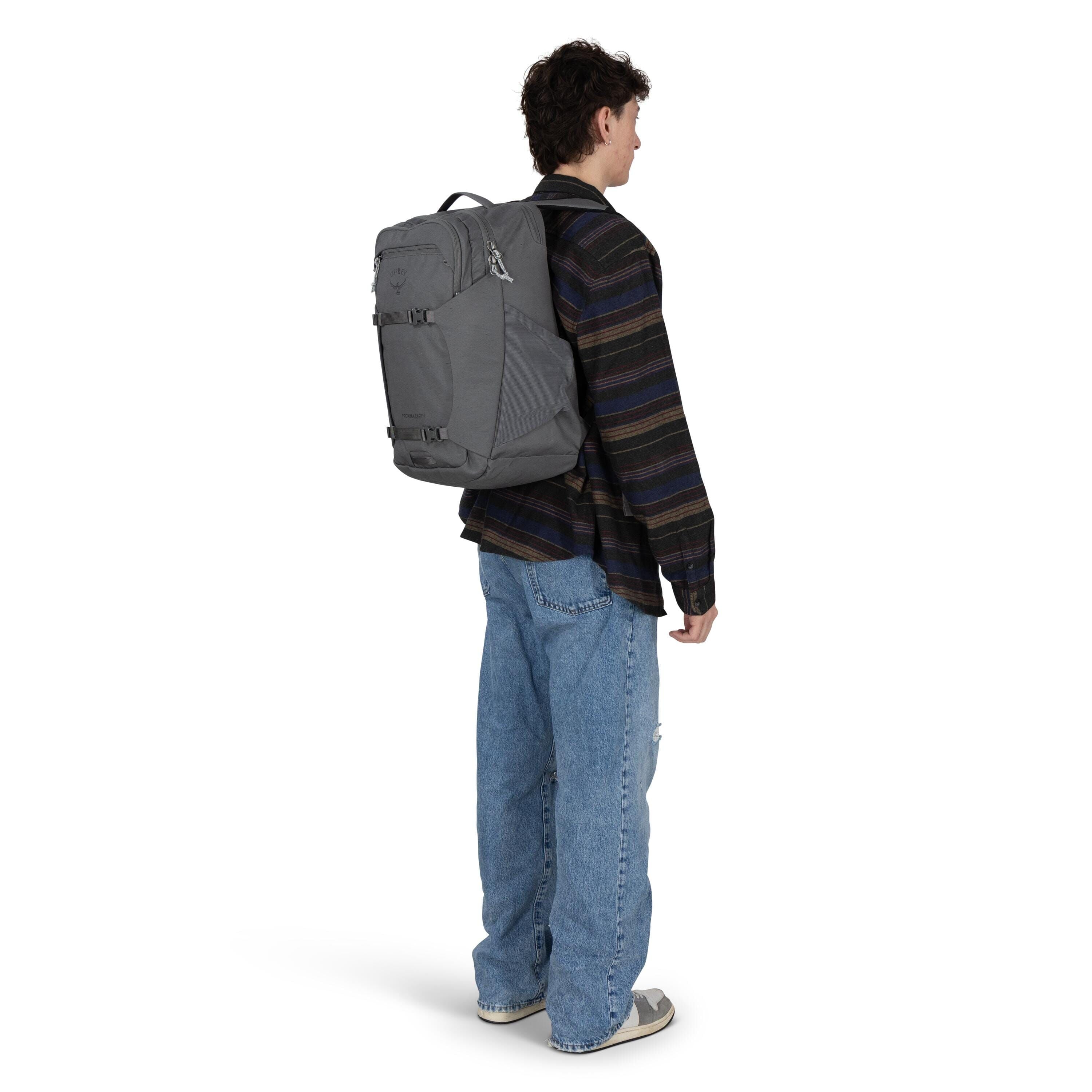 The image shows a person standing and facing slightly to the right, wearing a grey backpack on their back. The backpack features the Osprey logo and has multiple compartments and zippers, with side compression straps visible. The individual is dressed in