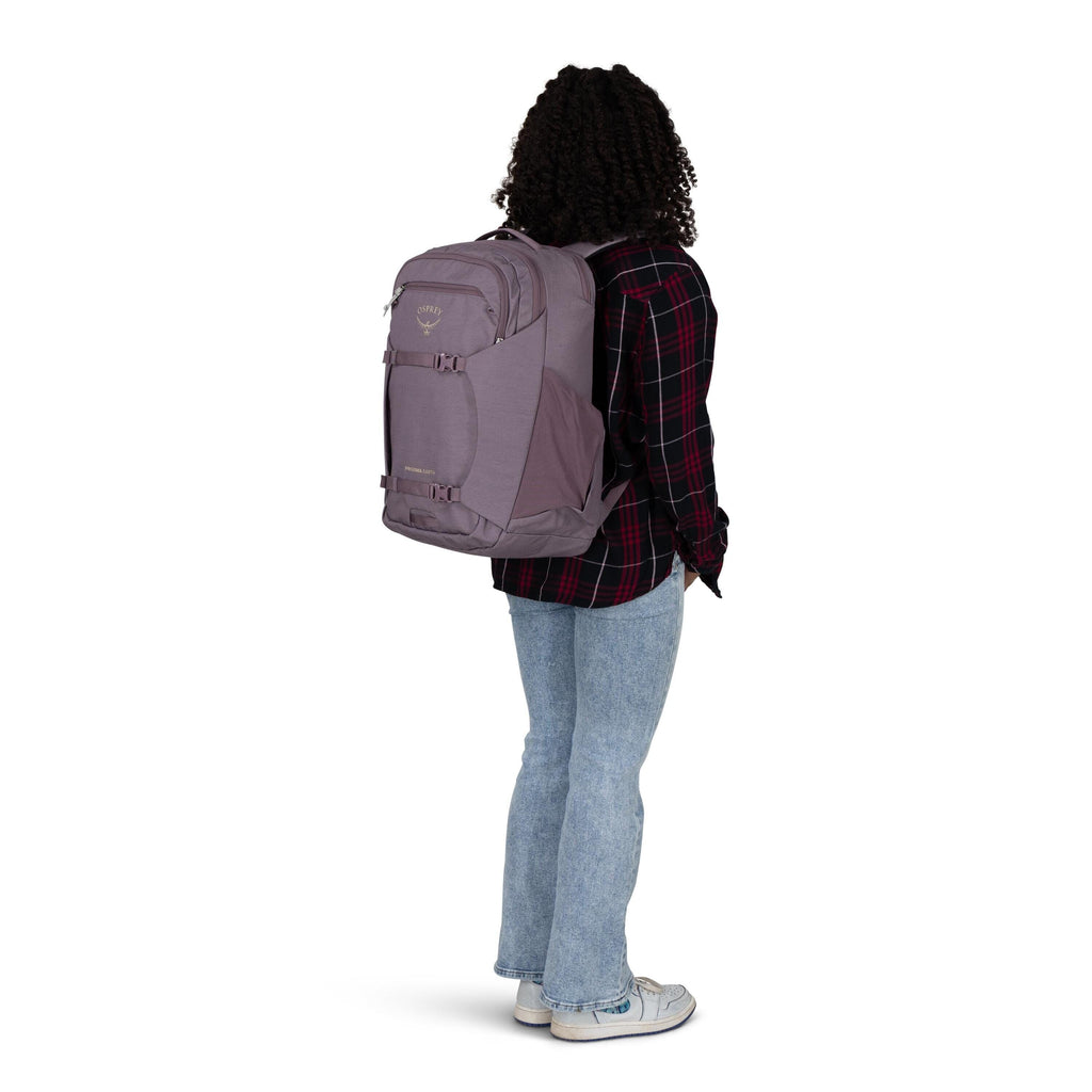The image shows a person standing and facing away from the camera, wearing a light purple Osprey backpack. The person is dressed in a black and red plaid shirt, light blue jeans, and white sneakers. Their hair is curly and shoulder-length. The scene is se