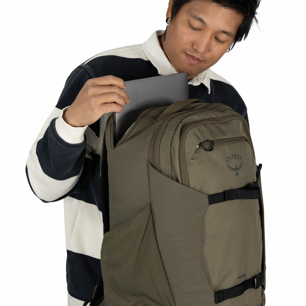 The image shows a person wearing a striped, long-sleeved shirt with wide black and white stripes, inserting a laptop into an Osprey backpack. The backpack is a neutral brown color and has the Osprey logo prominently displayed on the top front pocket. The