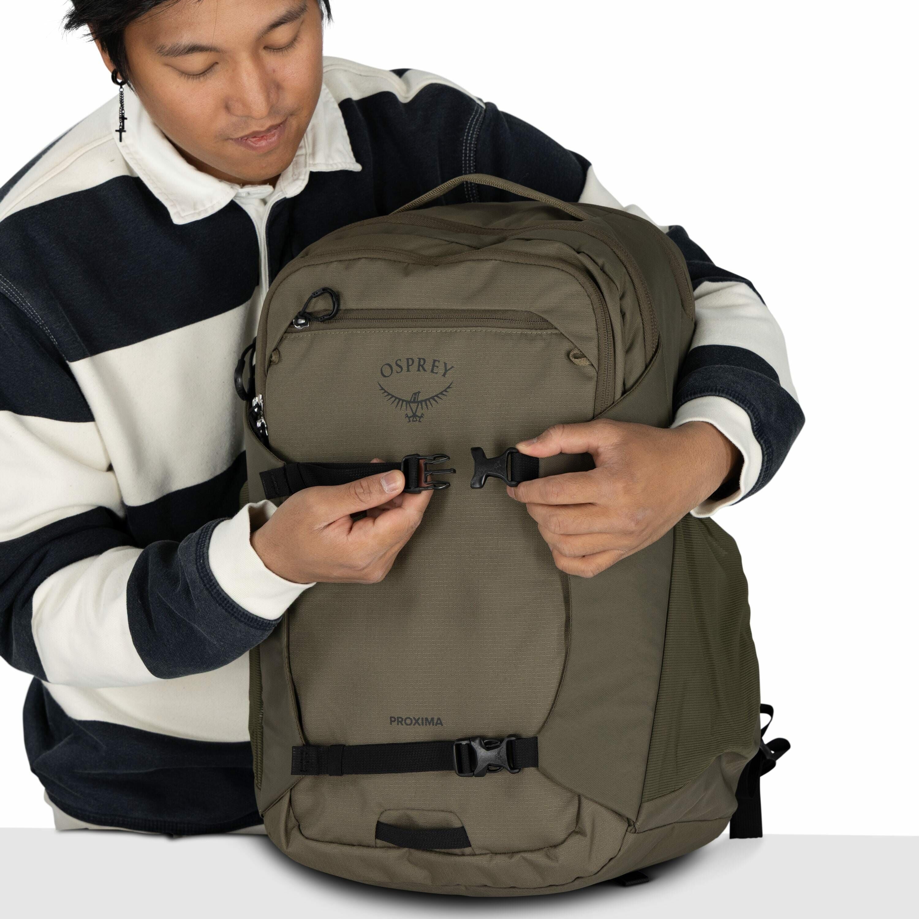 The image shows a person wearing a black and white striped sweater, holding a large backpack. The backpack is olive green and features the "Osprey" brand logo with an accompanying wing design prominently displayed on the front. The person is adjusting one