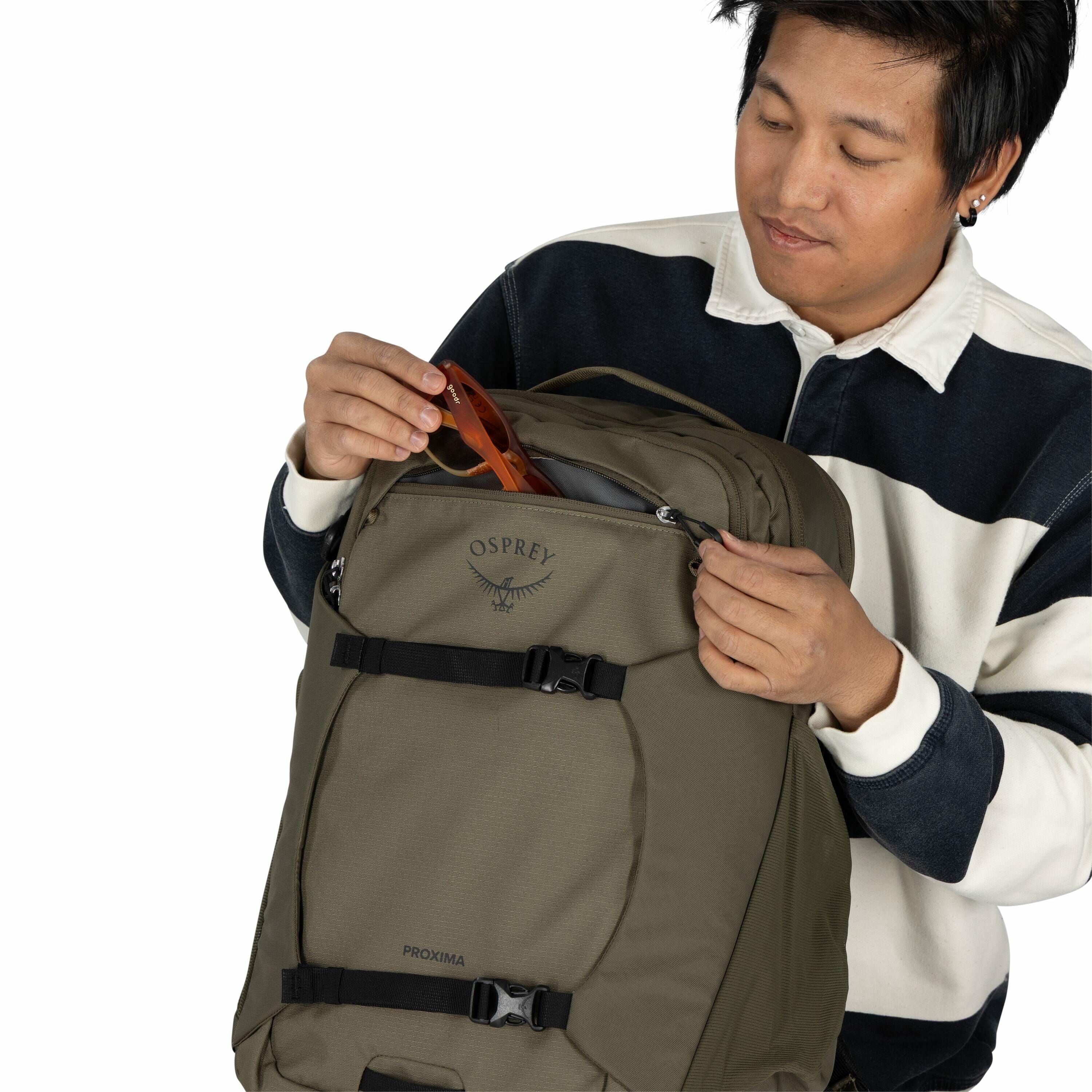 The image shows a person holding a khaki-colored Osprey backpack model "Proxima." The person is wearing a black and white striped long-sleeve shirt and is in the process of placing or retrieving a pair of brown sunglasses from the backpack's front pocket.