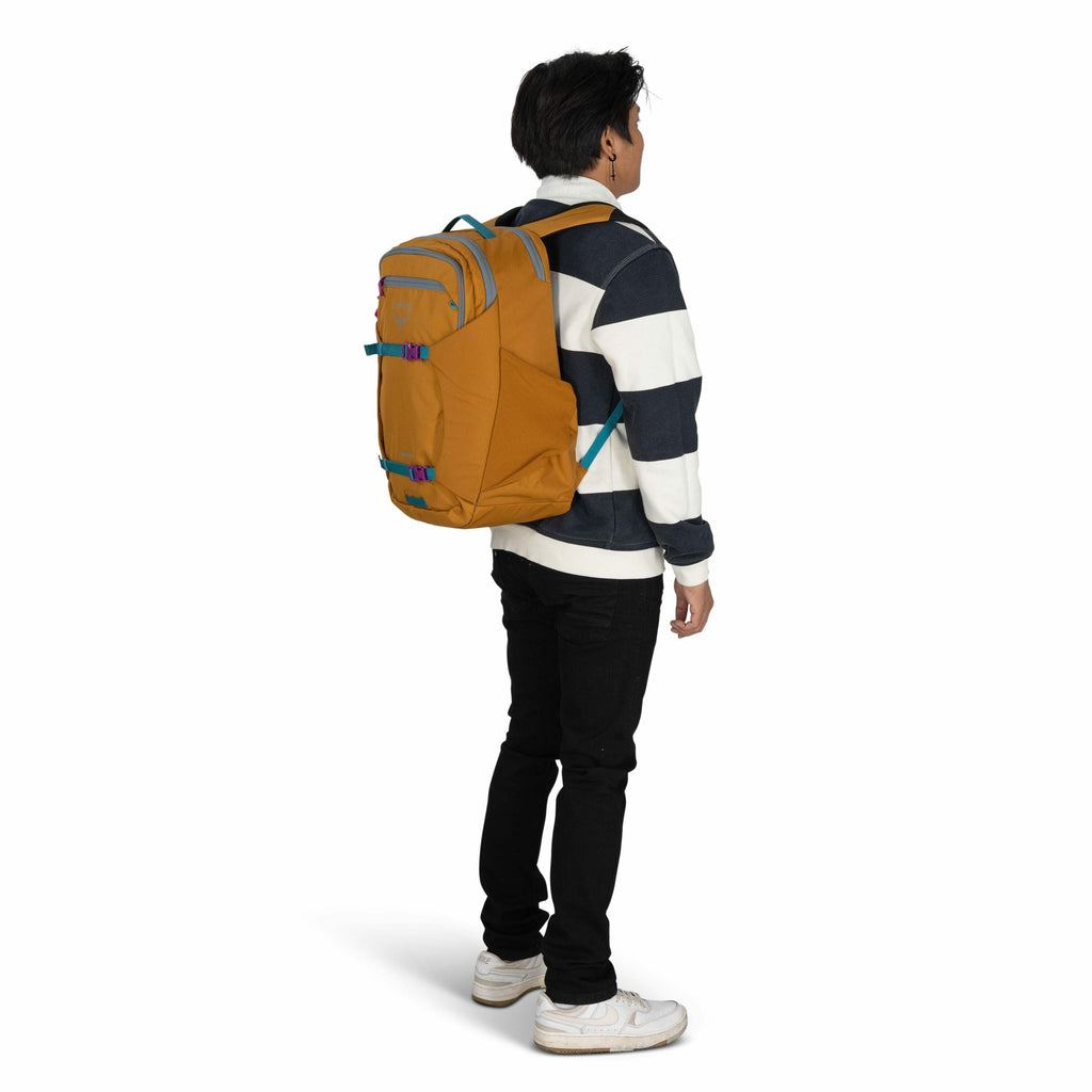 The image shows a person standing sideways, wearing a striped sweatshirt with thick navy and white horizontal stripes. They have a mustard-colored backpack with turquoise accents and straps, slung over their shoulders. The person is wearing black pants an