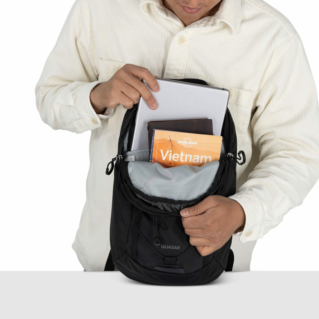 The image shows a man wearing a light-colored corduroy shirt, looking down as he opens a black backpack with the brand name "Quasar" on it. Inside the backpack, there are several items, including a tablet, a book with the title "Lonely Planet Vietnam," an