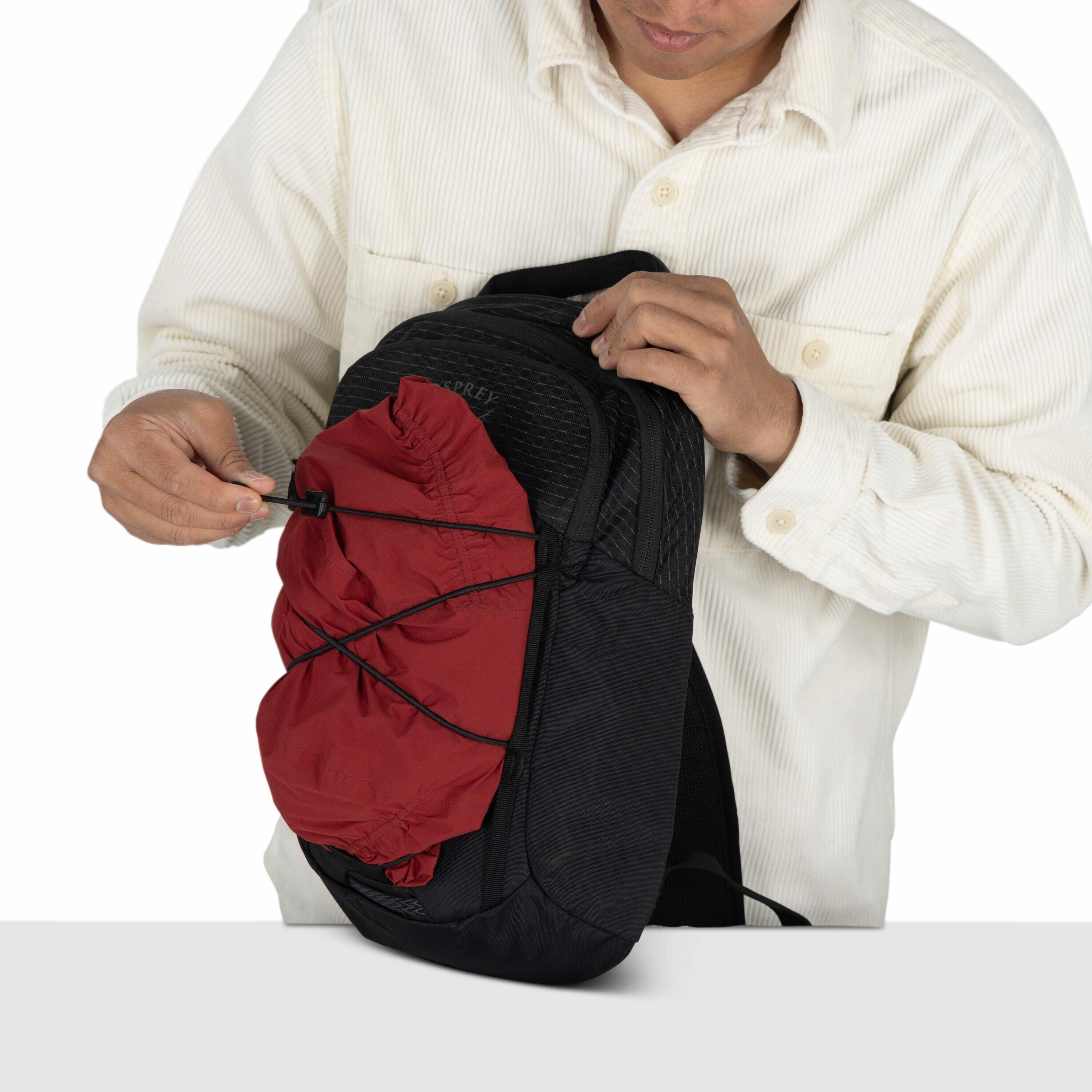 The image depicts a person in a light-colored, ribbed button-up shirt adjusting a black backpack. The individual is focused on securing a red item, possibly a jacket or bag cover, using the elastic bungee cord system on the front of the backpack. The back
