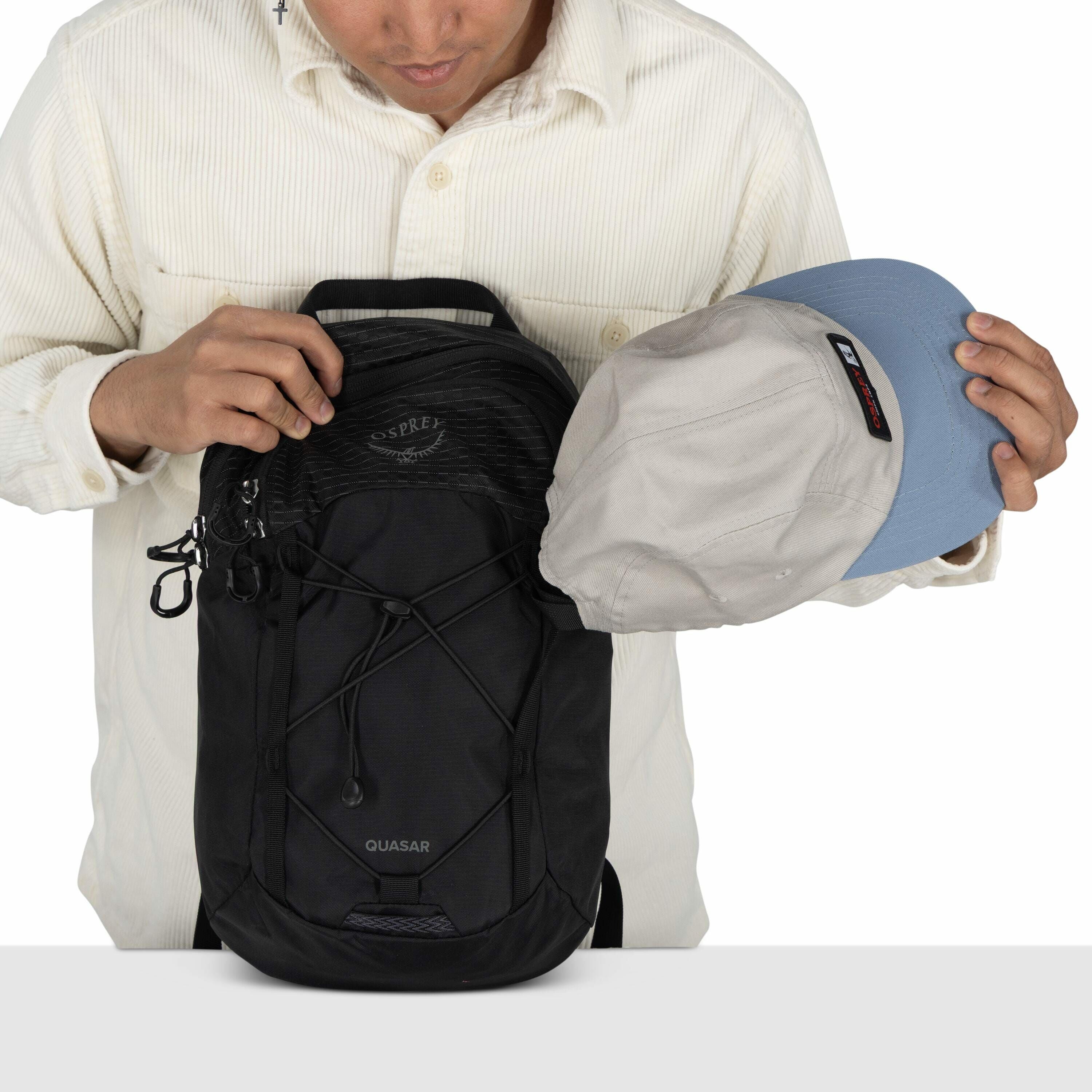In the image, a person wearing a light-colored, striped button-up shirt is seen placing a light gray and blue cap inside a black Osprey backpack. The backpack has "Quasar" labeled on the front and features a crisscross bungee cord on the exterior. The per