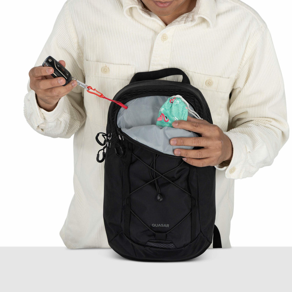 The image shows a person wearing a light-colored, corduroy shirt, interacting with a black Quasar backpack. The person is holding a set of keys attached to a red lanyard, which is clipped inside the backpack, suggesting an internal key attachment point. I