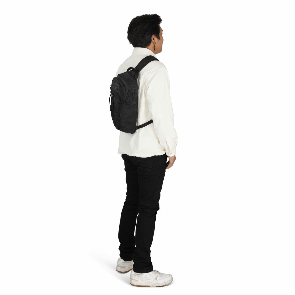 The image shows a man standing and facing slightly to the right, wearing a casual outfit. He is dressed in a white shirt, black jeans, and white sneakers. On his back, he carries a small black backpack with the Eagle Creek logo, which is secured with a wa