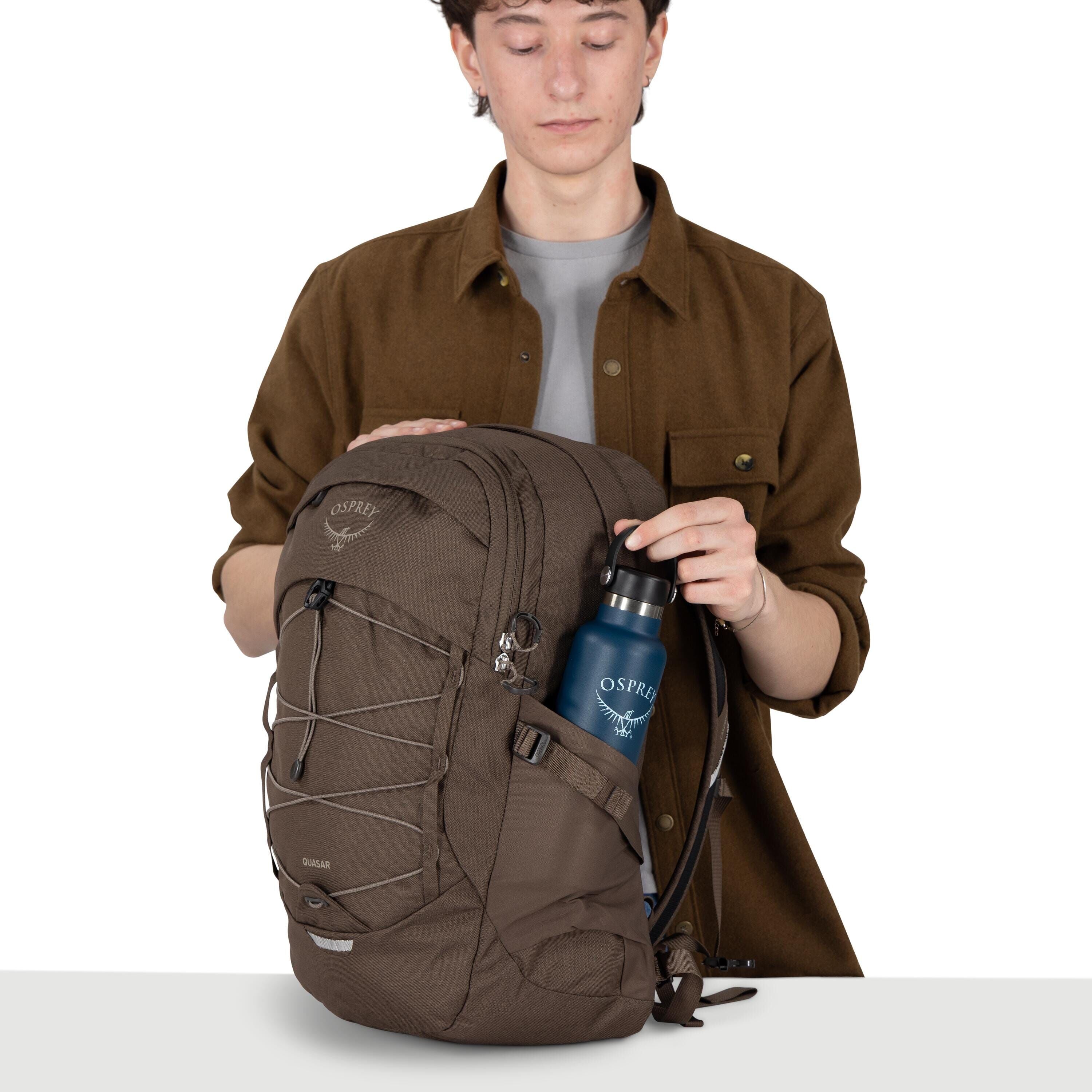 A person is holding a brown Osprey backpack on a white background. They are wearing a brown overshirt over a gray shirt, and their face is blurred. One hand rests on the top of the pack while the other places a blue Osprey-branded water bottle into a side