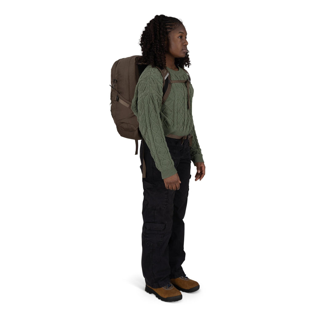 A person is standing in a three-quarter side view wearing outdoor clothing and a large backpack. They have on a textured green cable-knit sweater, black cargo pants, and brown hiking boots with rugged soles. The backpack is taupe with compression straps a