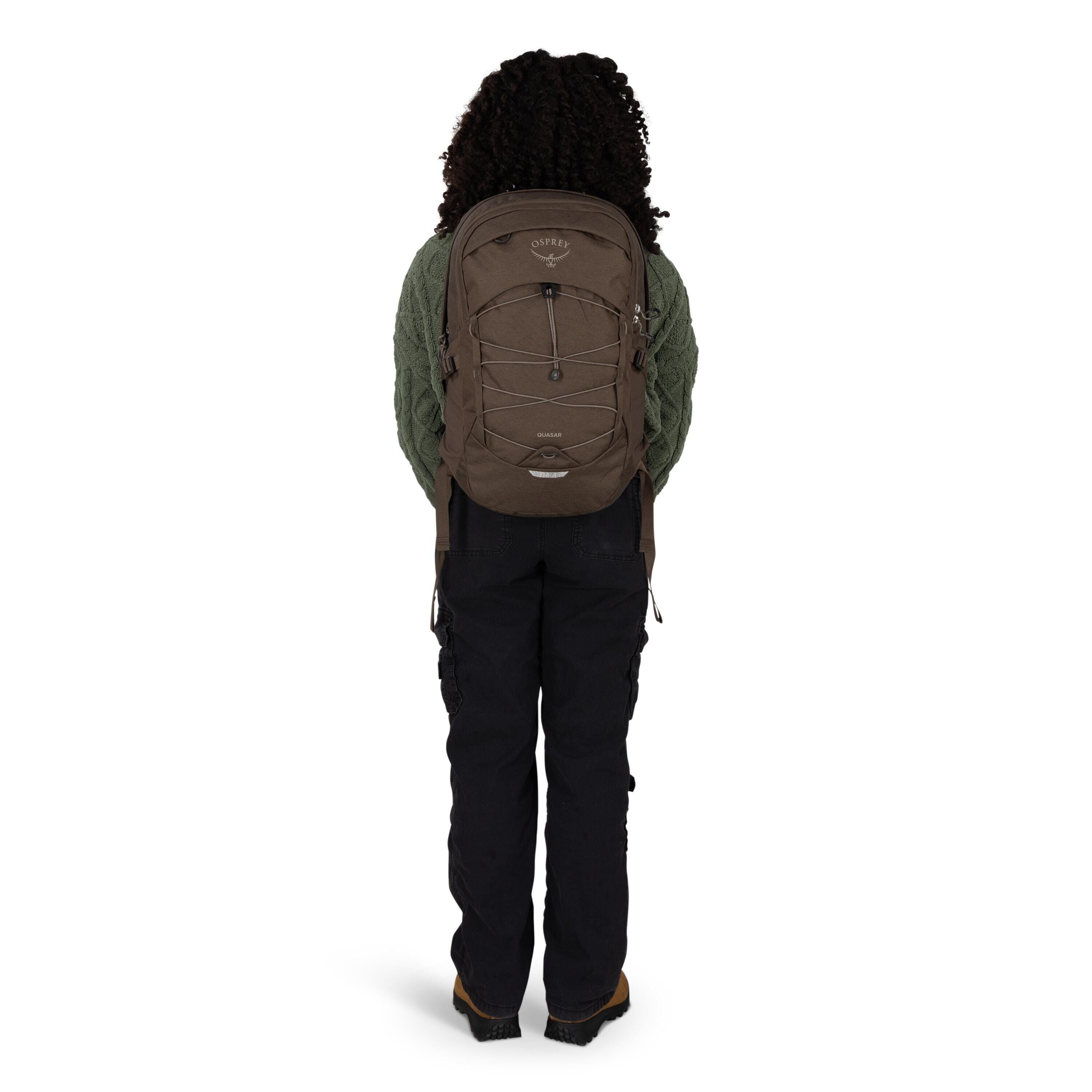 A person is standing facing away from the camera, wearing a brown Osprey backpack. The backpack has a front bungee cord system, side compression straps, and a small top pocket. They are dressed in a textured green sweater, black cargo-style pants, and bro