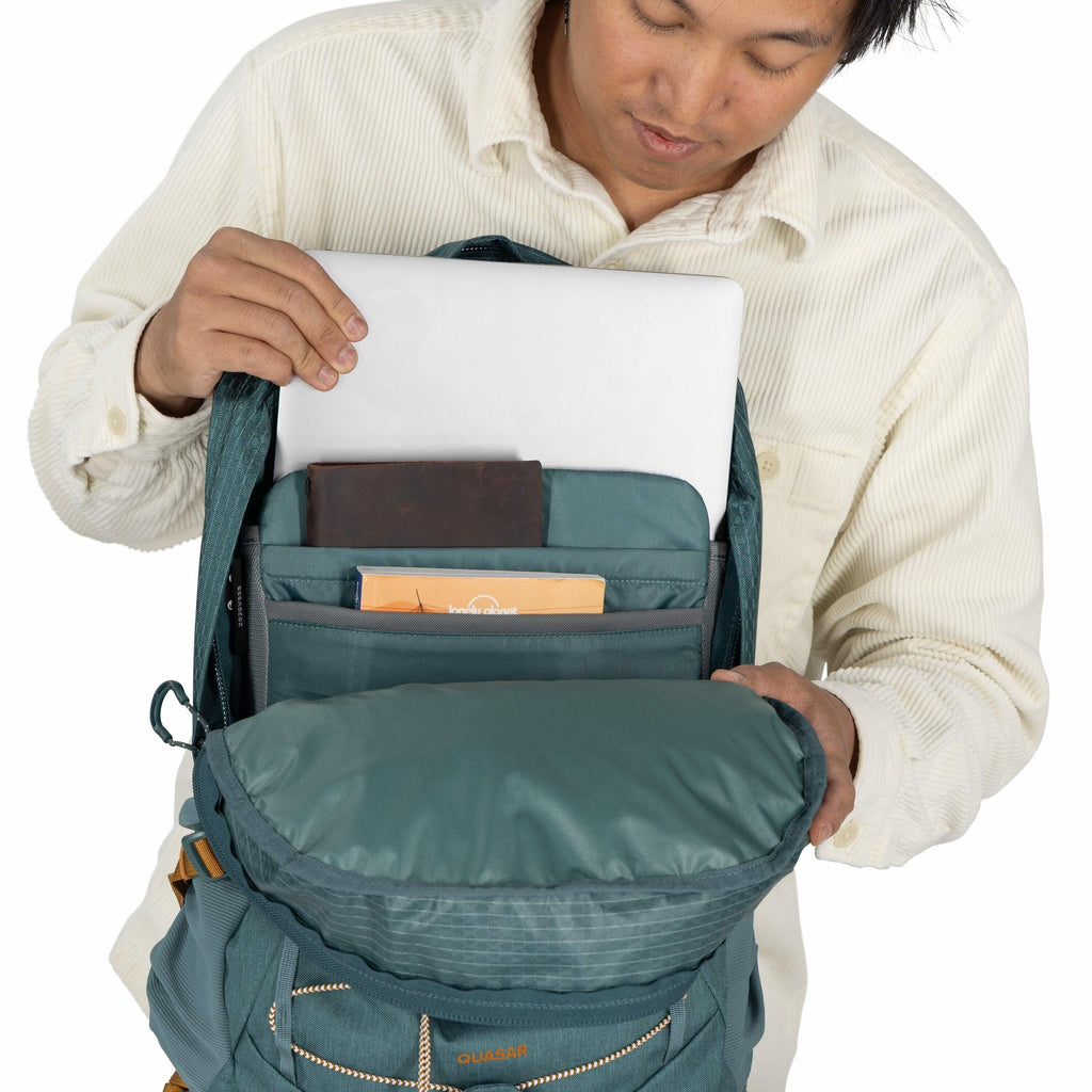 The image depicts a person wearing a cream-colored, corduroy shirt, opening a teal Quasar brand backpack to reveal its contents. Inside the backpack, a laptop is seen being pulled out from a dedicated laptop sleeve. The backpack also contains several item