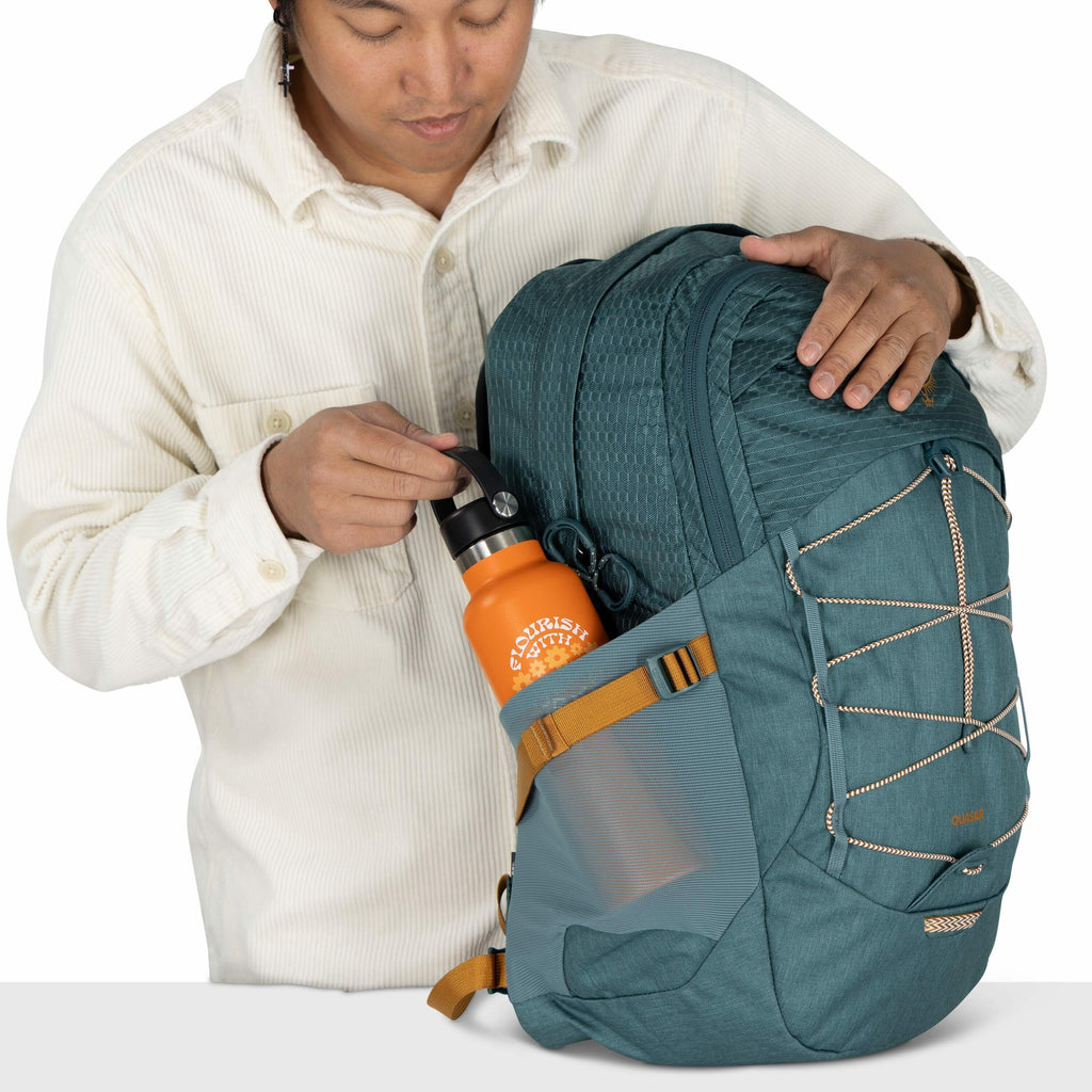 The image shows a person wearing a cream-colored corduroy button-up shirt, holding a teal-colored backpack. The backpack has a mesh side pocket where an orange water bottle labeled "Nourish with Nature" is being placed. The backpack features orange accent